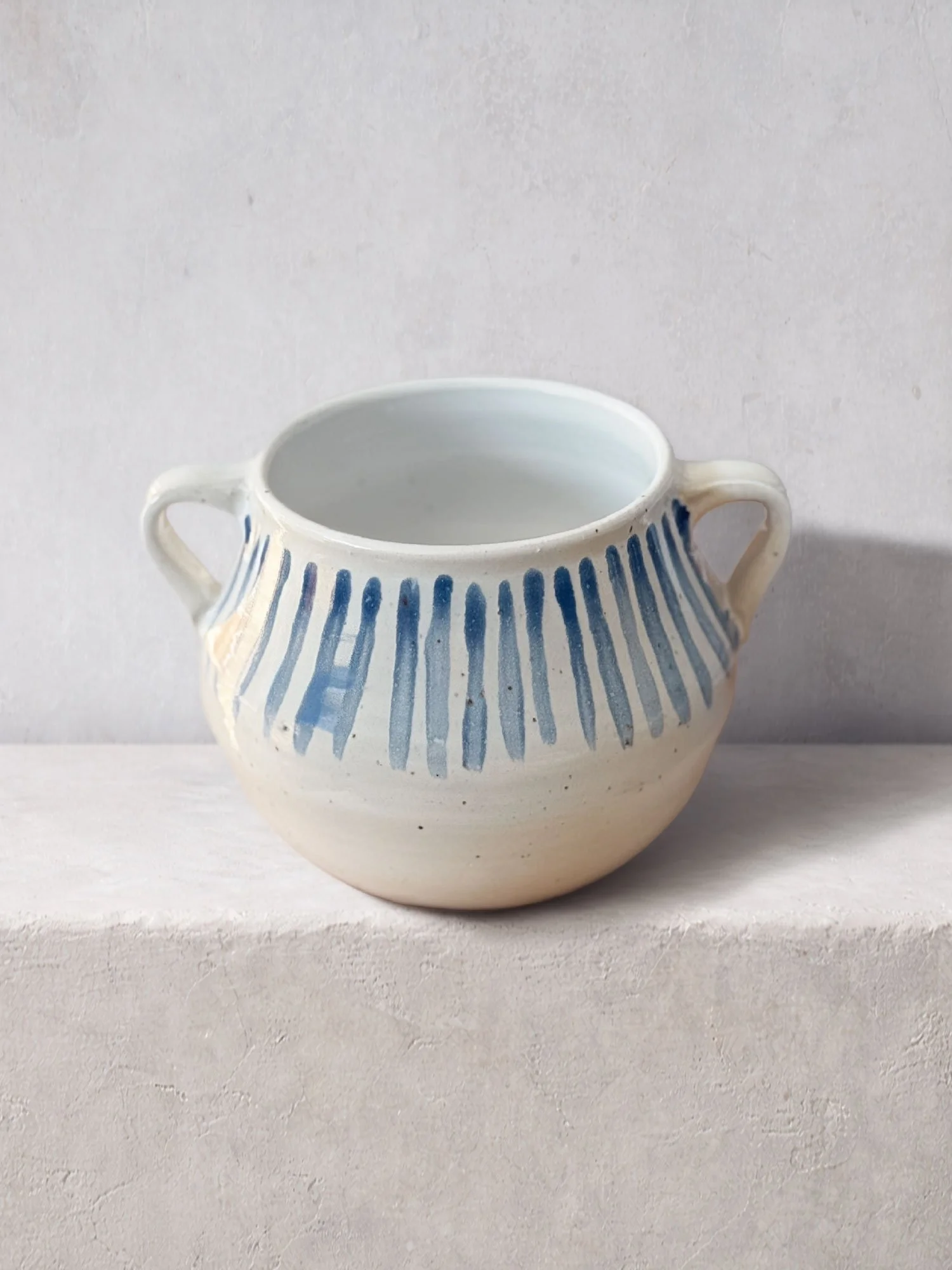 Handmade ceramic bowl with two handles, decorated with blue vertical markings on a warm white clay body.