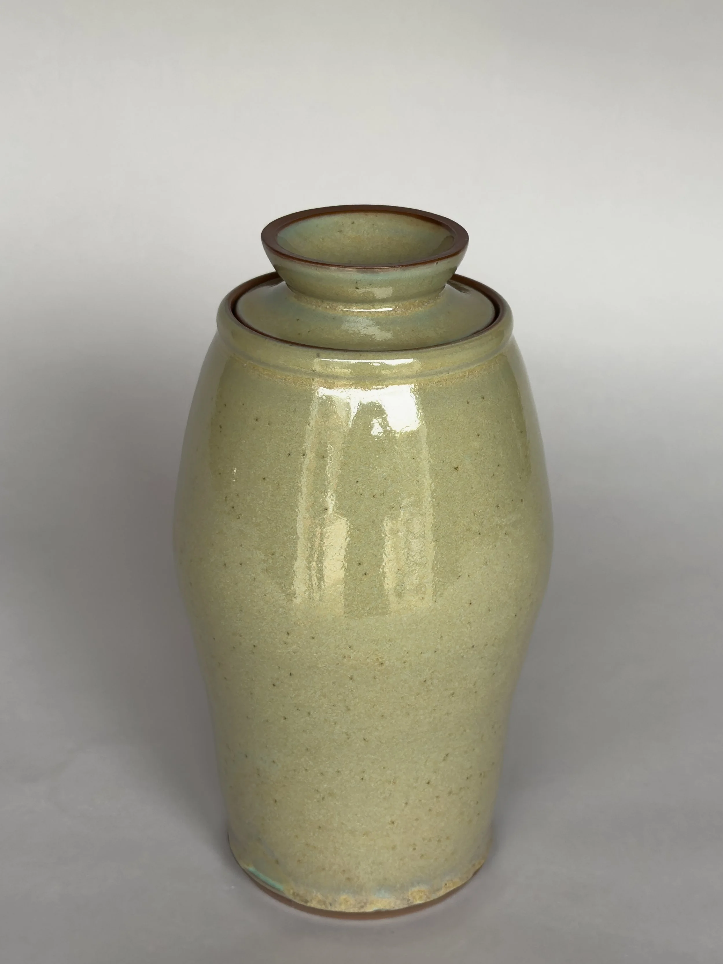 Tall handmade ceramic vase with lid and soft speckled sage green glaze by Jeanette Burgett Ceramics on a neutral gray background.