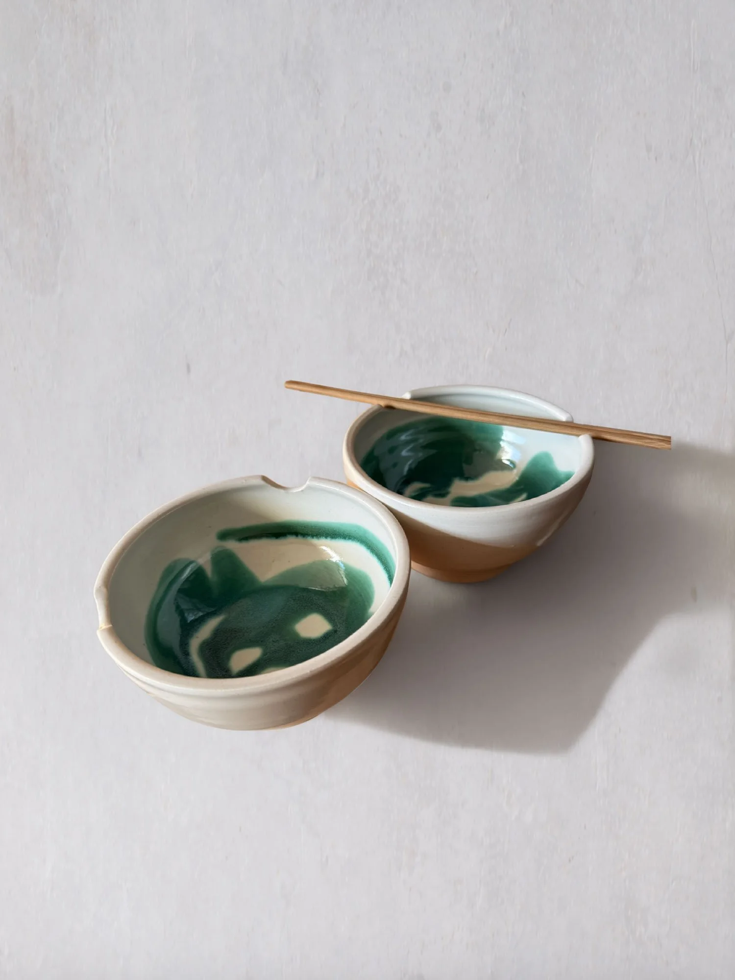 Two handmade ceramic bowls with green glazed interiors and natural clay exteriors, styled with wooden chopsticks on a textured surface.