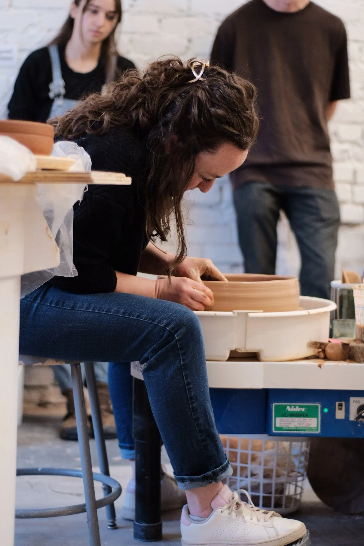 A woman shaping a clay pottery on a pottery wheel, with a woman and a man observing in the background.