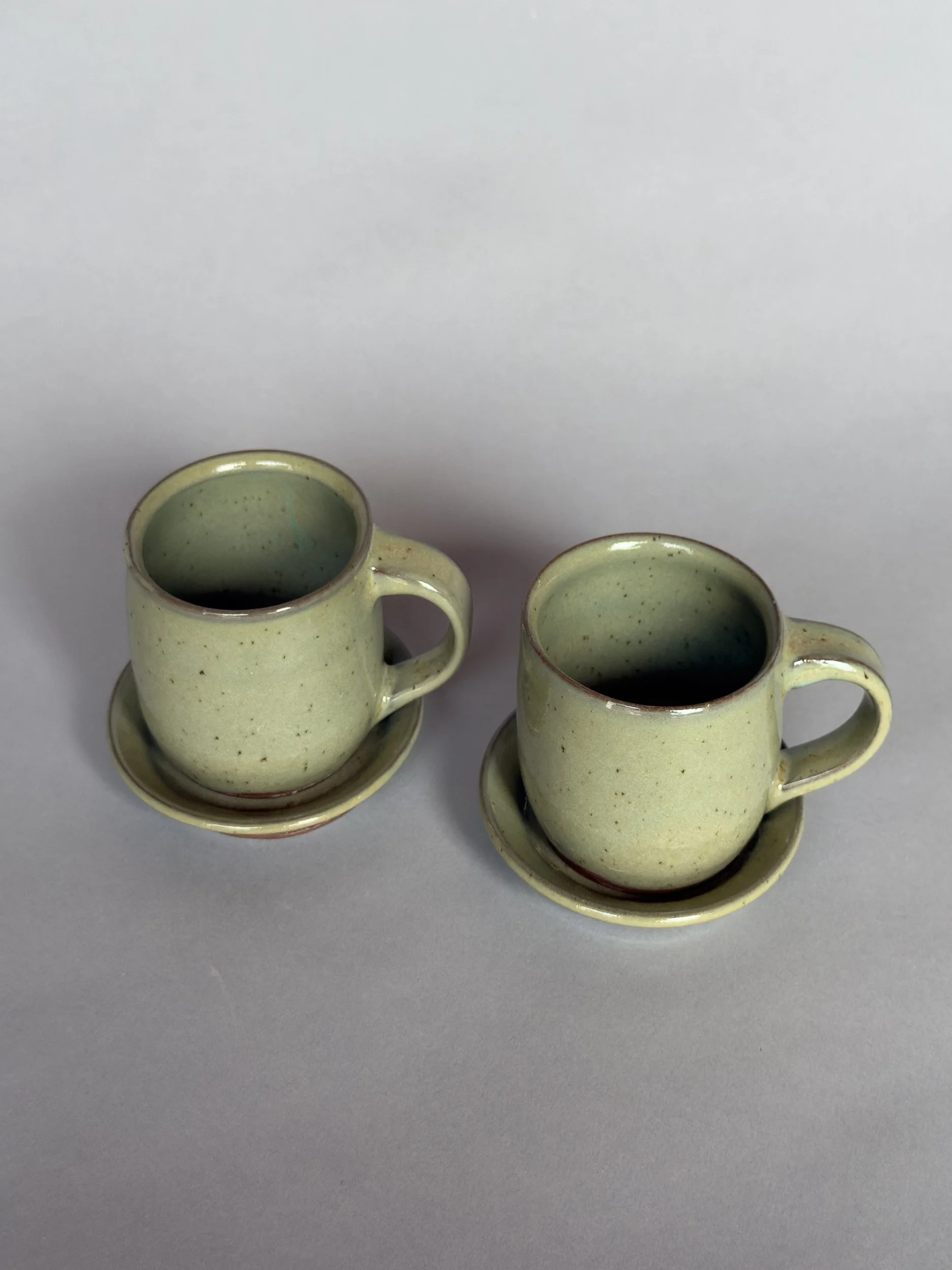 Two handmade ceramic espresso cups with matching saucers and soft speckled sage green glaze by Jeanette Burgett Ceramics on a neutral gray background.