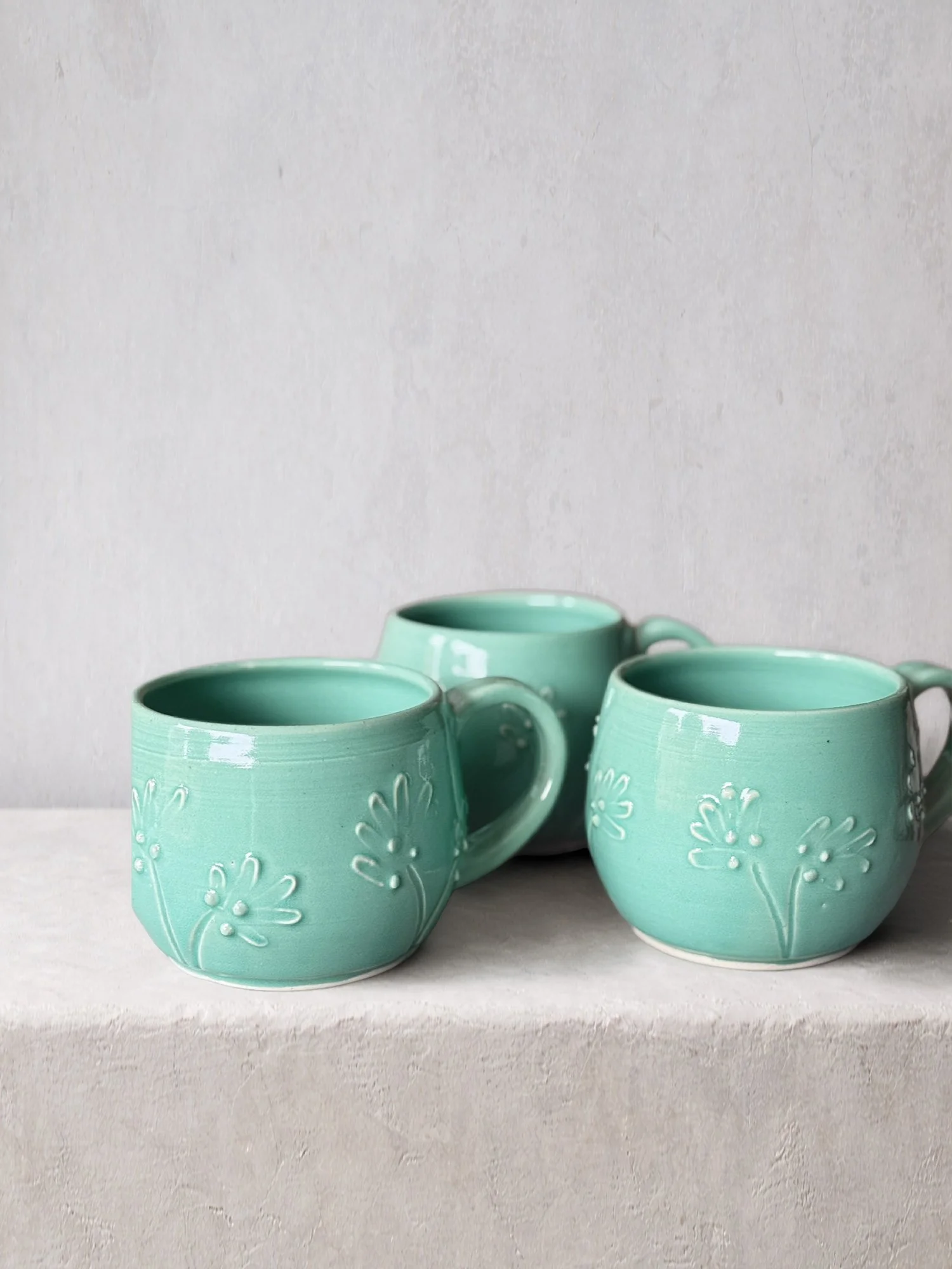 Three handmade ceramic mugs in a soft teal glaze with raised floral details, arranged on a neutral studio background.