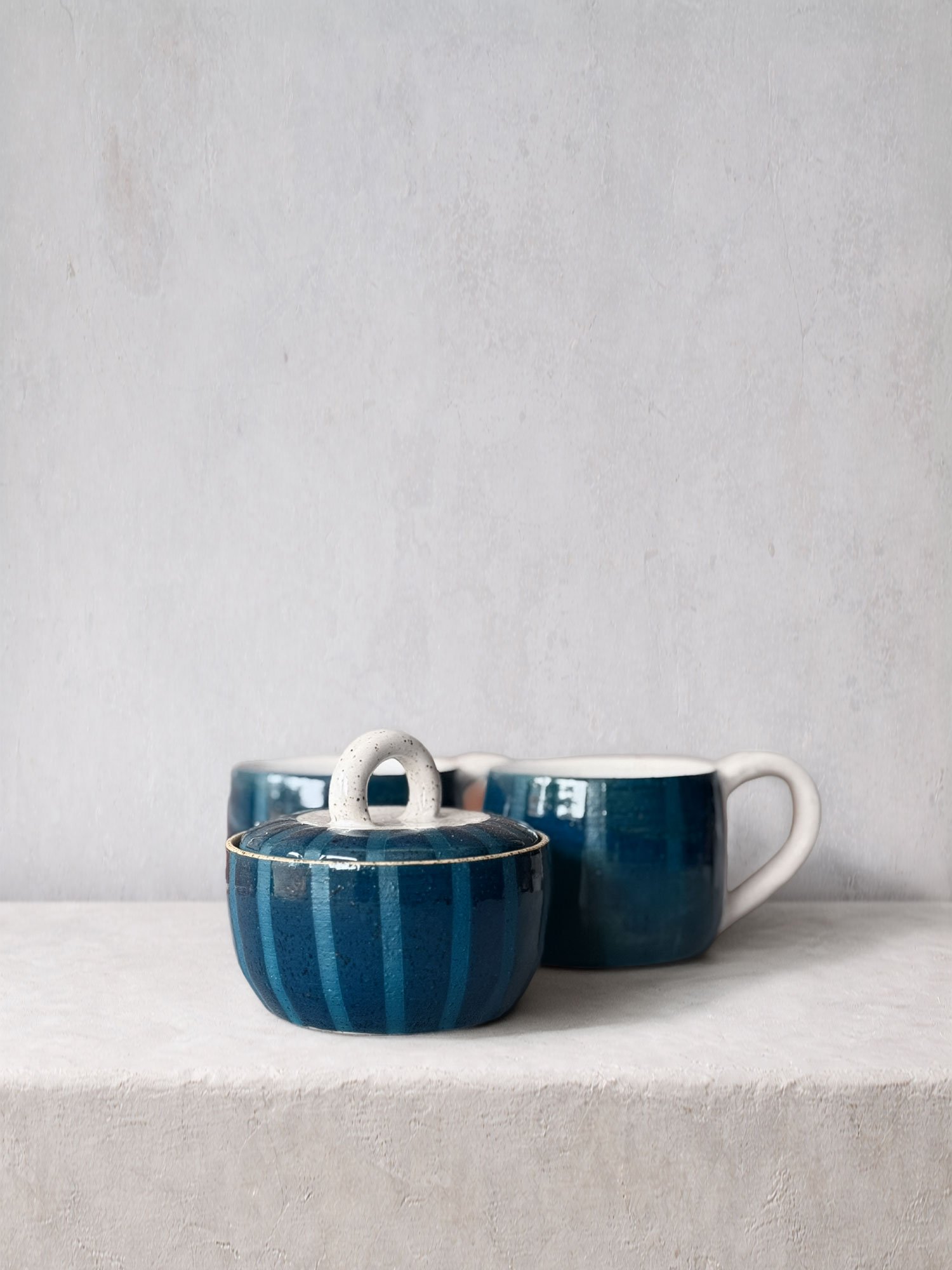 A set of three blue ceramic cups with white handles and a matching sugar bowl with a lid, displayed on a white surface against a light grey background.