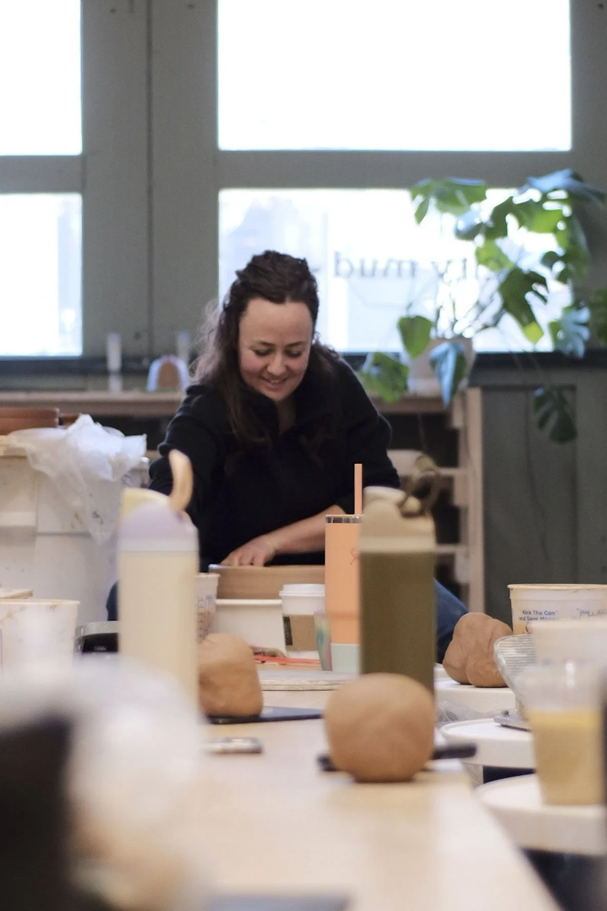 A woman with dark hair and a black shirt is smiling and working at a table surrounded by various items including paper cups, a pink water bottle, and a paper bag, in a bright room with large windows and a green plant.