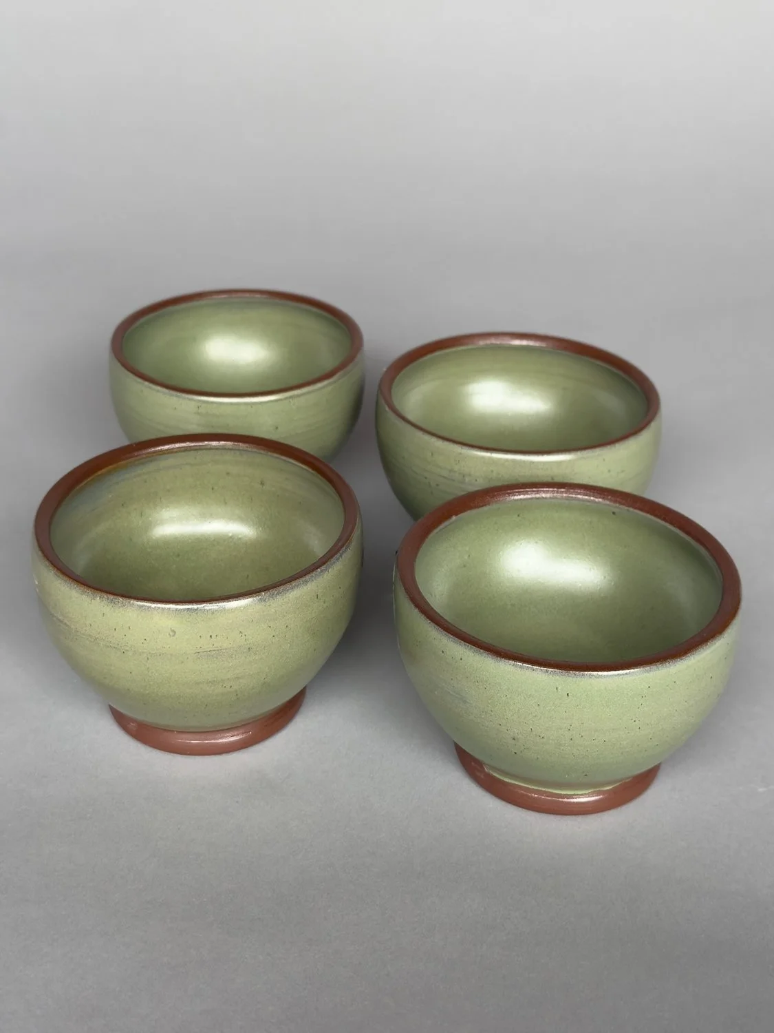 Set of four handmade ceramic bowls with soft sage green glaze and terracotta rims by Jeanette Burgett Ceramics on a neutral gray background.