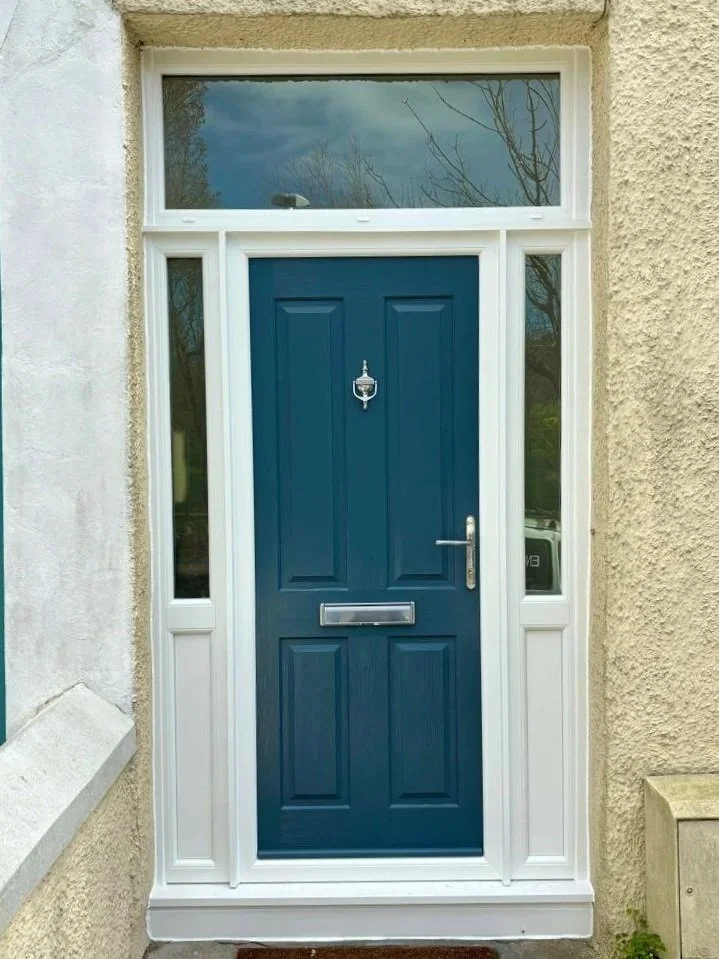 Blue front door with silver handle, letterbox, and door knocker, surrounded by white trim, with a transom window above and sidelights on each side, set in a yellow textured exterior wall.