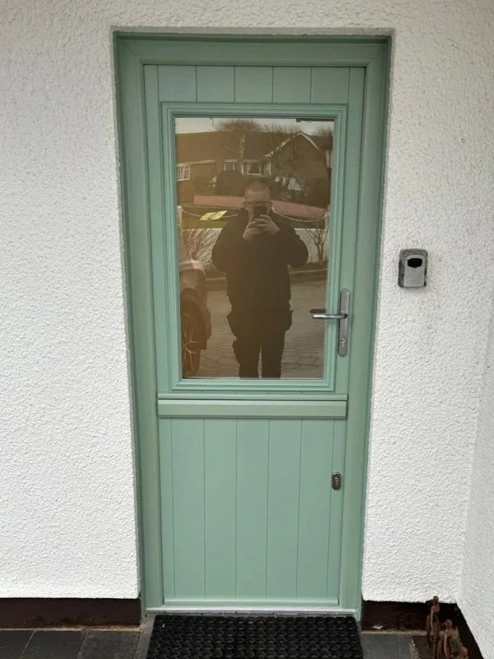 A green door with a glass window reflecting a person taking a photo outside, and a street scene behind them.