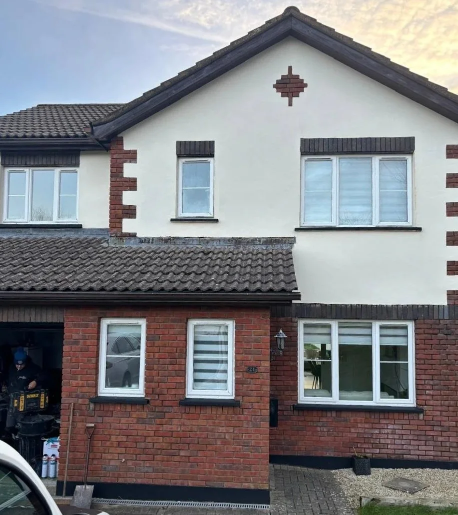 The upper part of a house with a white facade, small windows, and decorative brick accents. The lower part is made of red bricks with larger windows, a lamp post, and some tools near the garage. The roof has brown shingles.