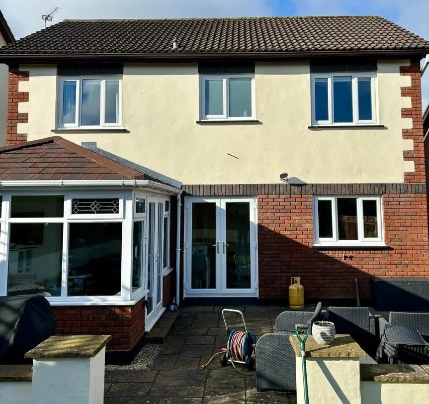 Backyard of a two-story house with a brick and stucco exterior, multiple windows, and a patio area with outdoor furniture, gardening tools, and a gas cylinder.
