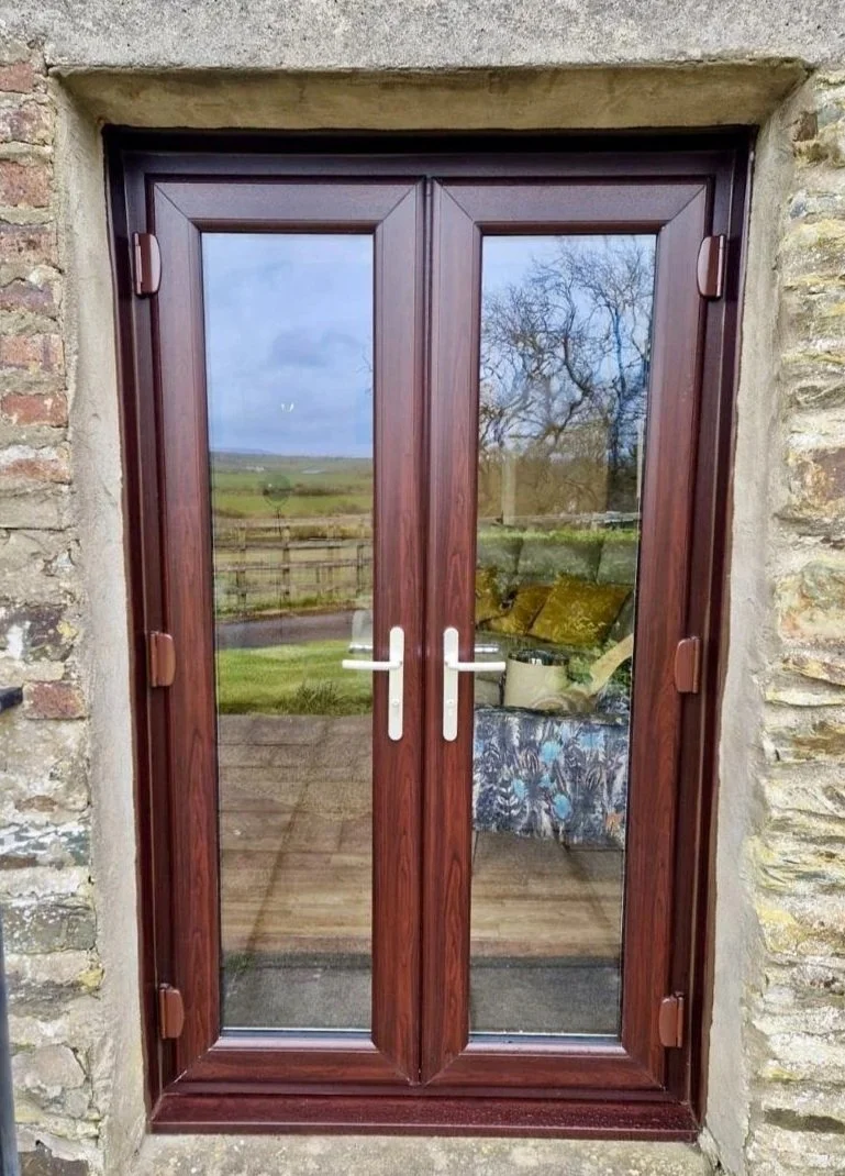 Double glass doors with wooden frames, installed in a stone wall, reflecting an outdoor landscape and a room interior with furniture.