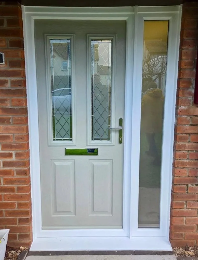 A white front door with two tall glass panels and a mailbox slot, flanked by brick walls and a side window, with a reflection of a person and some outdoor scenery in the glass.