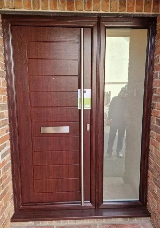Front door with a wooden sliding door and a glass panel, brick exterior wall, and a person's reflection visible in the glass.