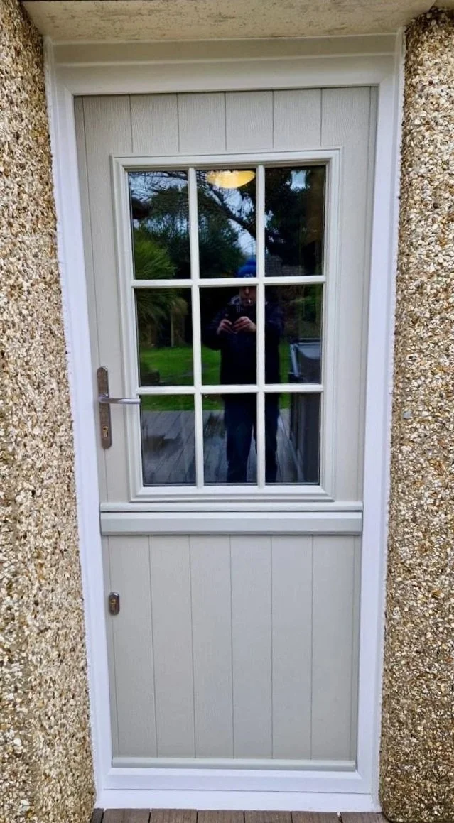 Exterior view of a door with a large glass window divided into small panes, set between textured concrete walls, with a reflection of the person taking the photo visible in the glass.