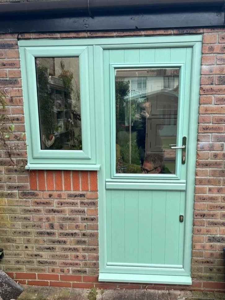 A brick house exterior with a mint green door that has a side window. The door has a glass panel and a handle, and there is an adjoining window to the left of the door.