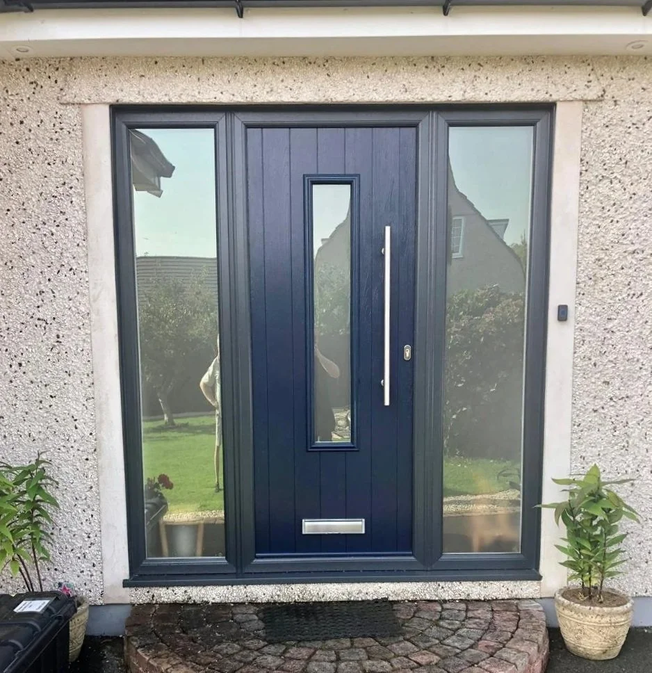 Front door with dark blue panel, vertical window, silver handle, mail slot, flanked by glass side panels, on a brick pathway, with potted plants on each side.