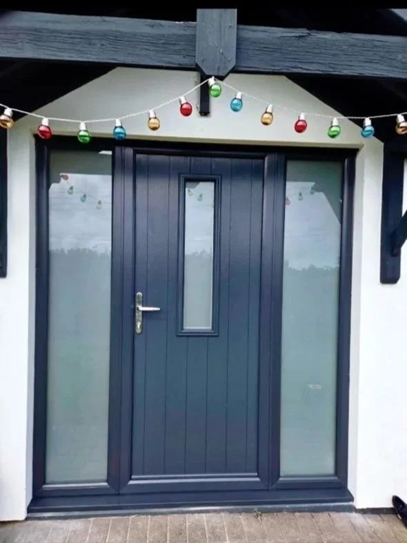 Front door with dark blue frame, small vertical window in the middle, decorated with colorful string lights overhead, outside area with brick pavement.