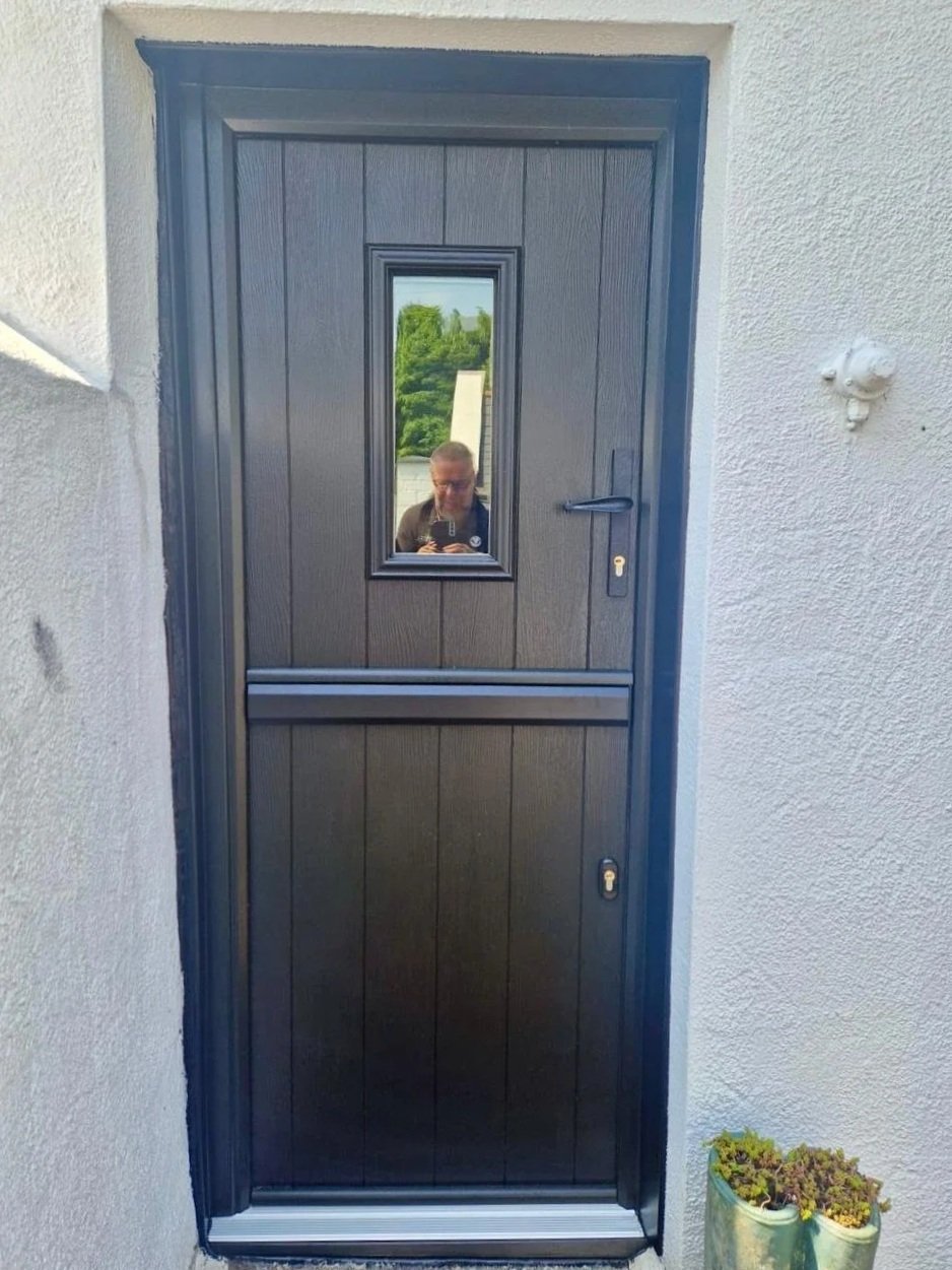 Black exterior door with a narrow rectangular window in the upper half, set in a white stucco wall. The door has a handle and keyhole, with a small plant in the bottom right corner.