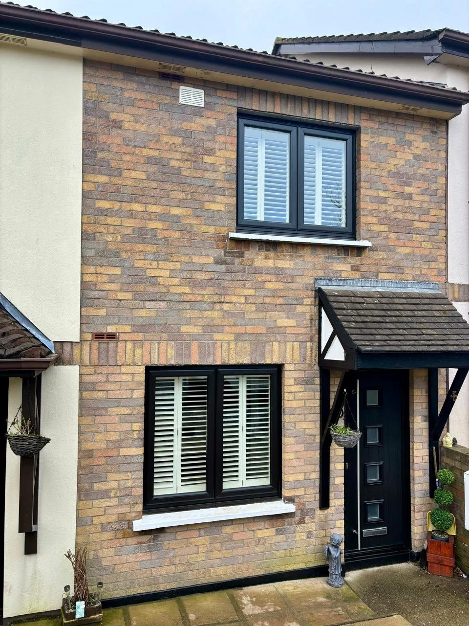A two-story brick house with black-framed windows with white shutters, a black front door with decorative panels, and a small porch with hanging baskets and potted plants.