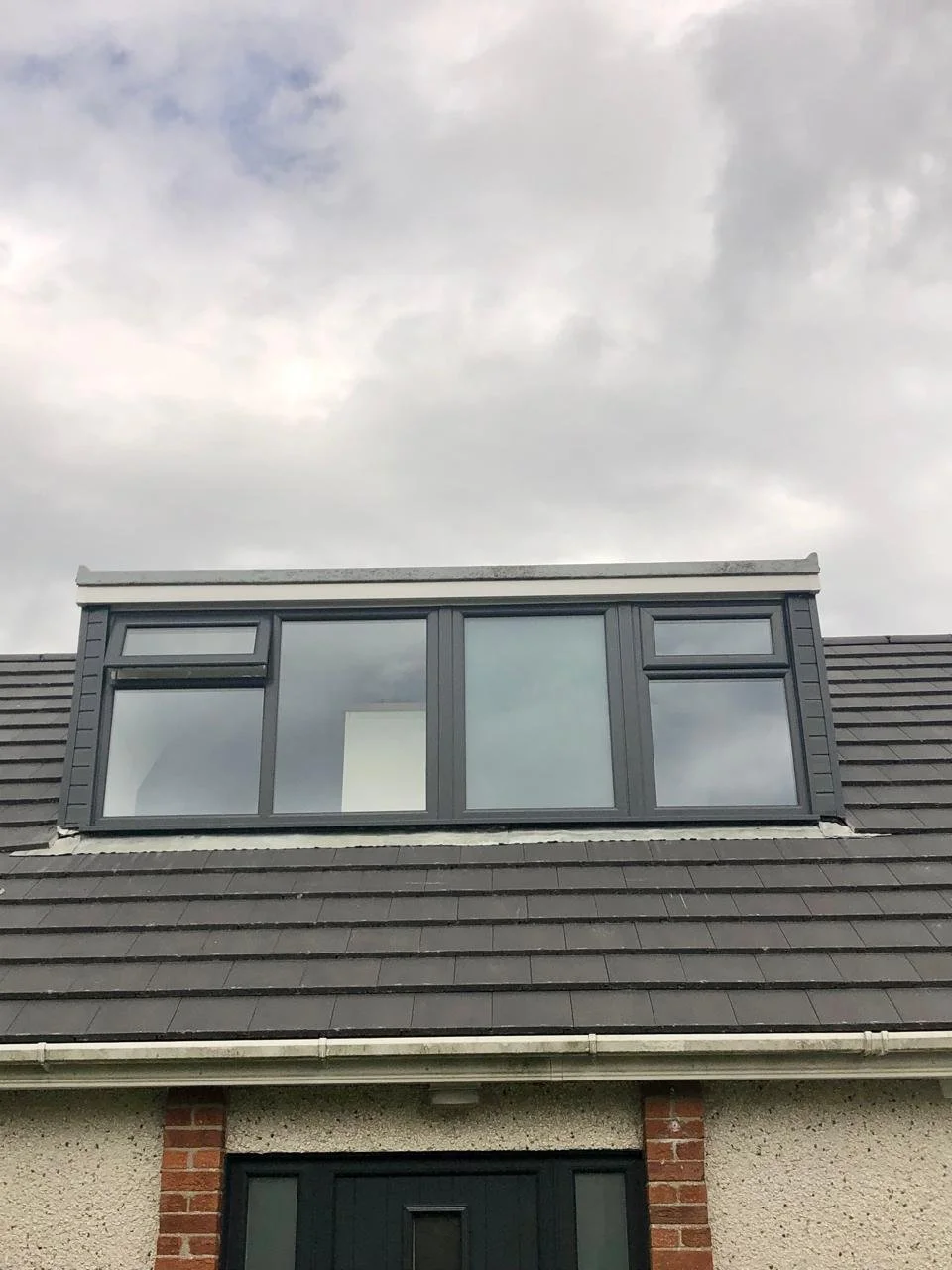 Attic dormer with three large windows on a sloped roof, grey frames, under cloudy sky.