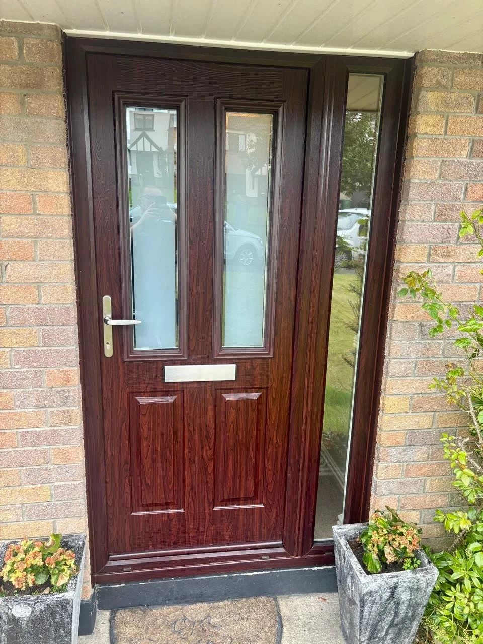 Front door of a house with a dark wood finish, vertical glass panels, and a silver mail slot, flanked by brick walls and potted plants.