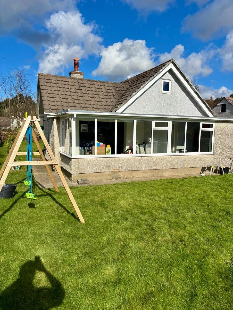 A house with a porch or sunroom, a grassy yard, a wooden swing set, and a bright blue sky with clouds.
