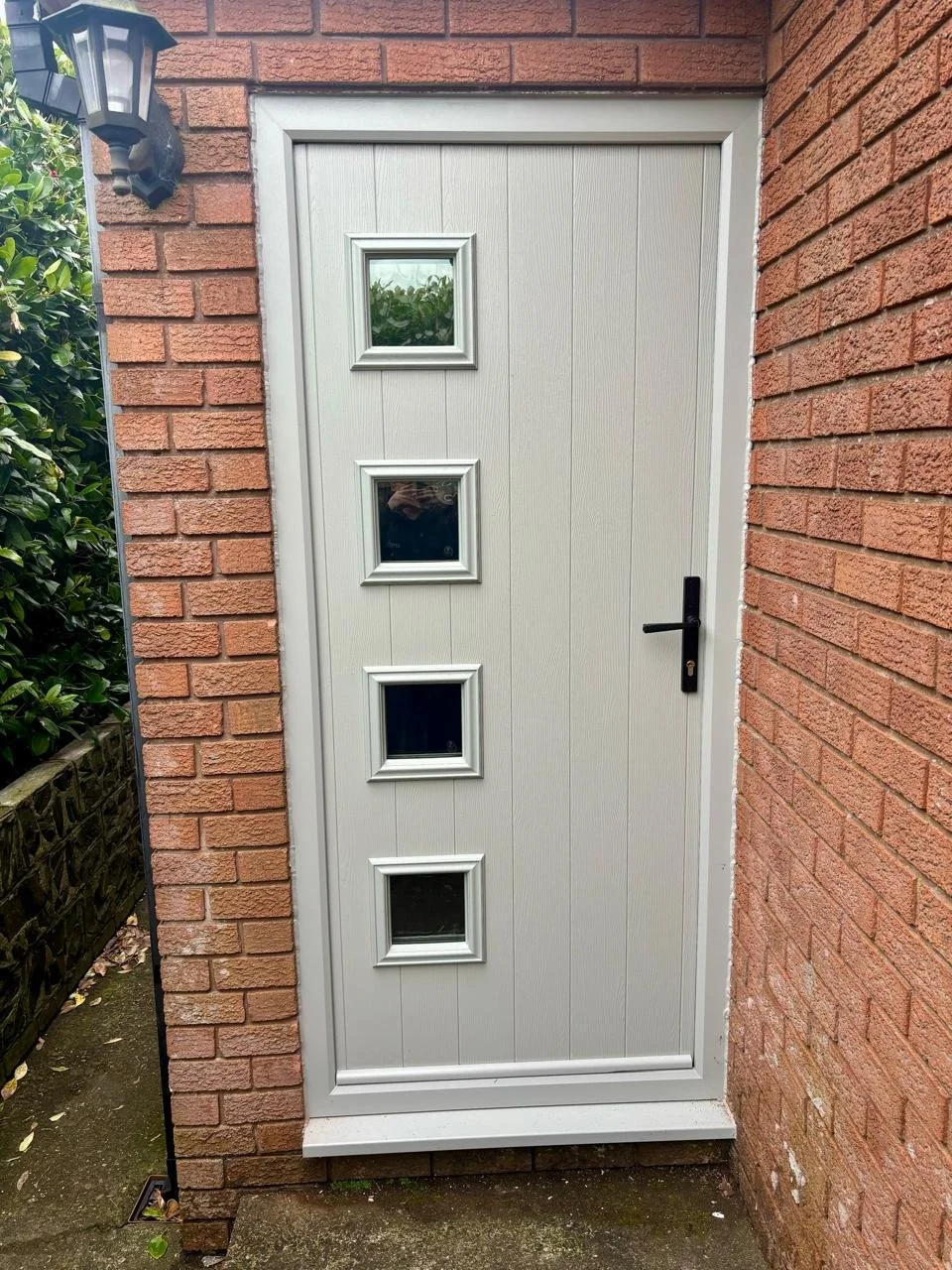 A white exterior door with four small square windows aligned vertically, set in brick walls, with a black handle and lock, and an outdoor lantern above on the left.