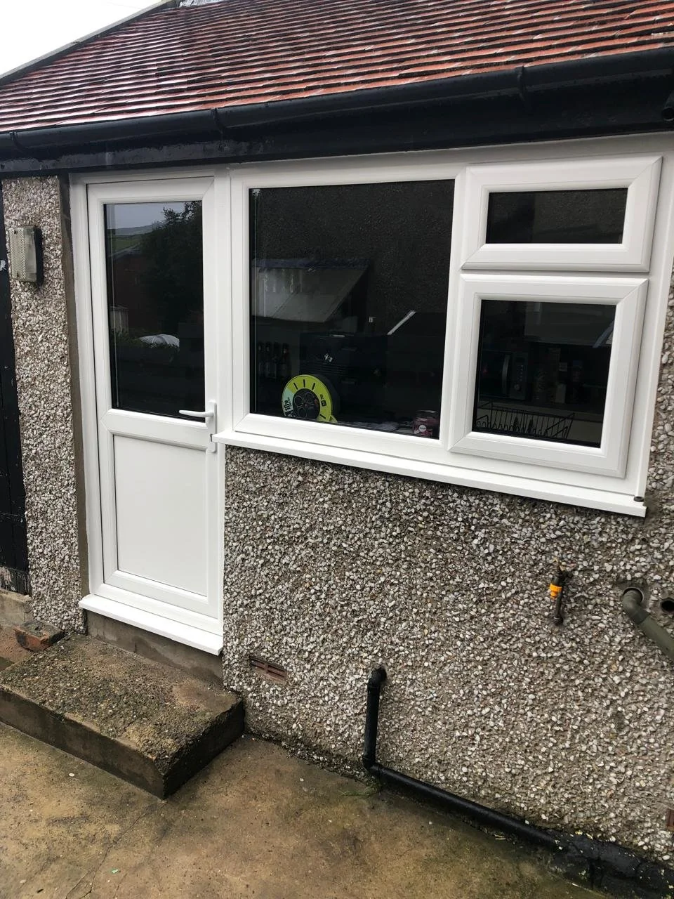 Exterior of a house with a gravel-textured wall, a white-framed front door, and a window with a white frame and two smaller windows attached. There is a small concrete step outside the door and some visible pipes on the wall.