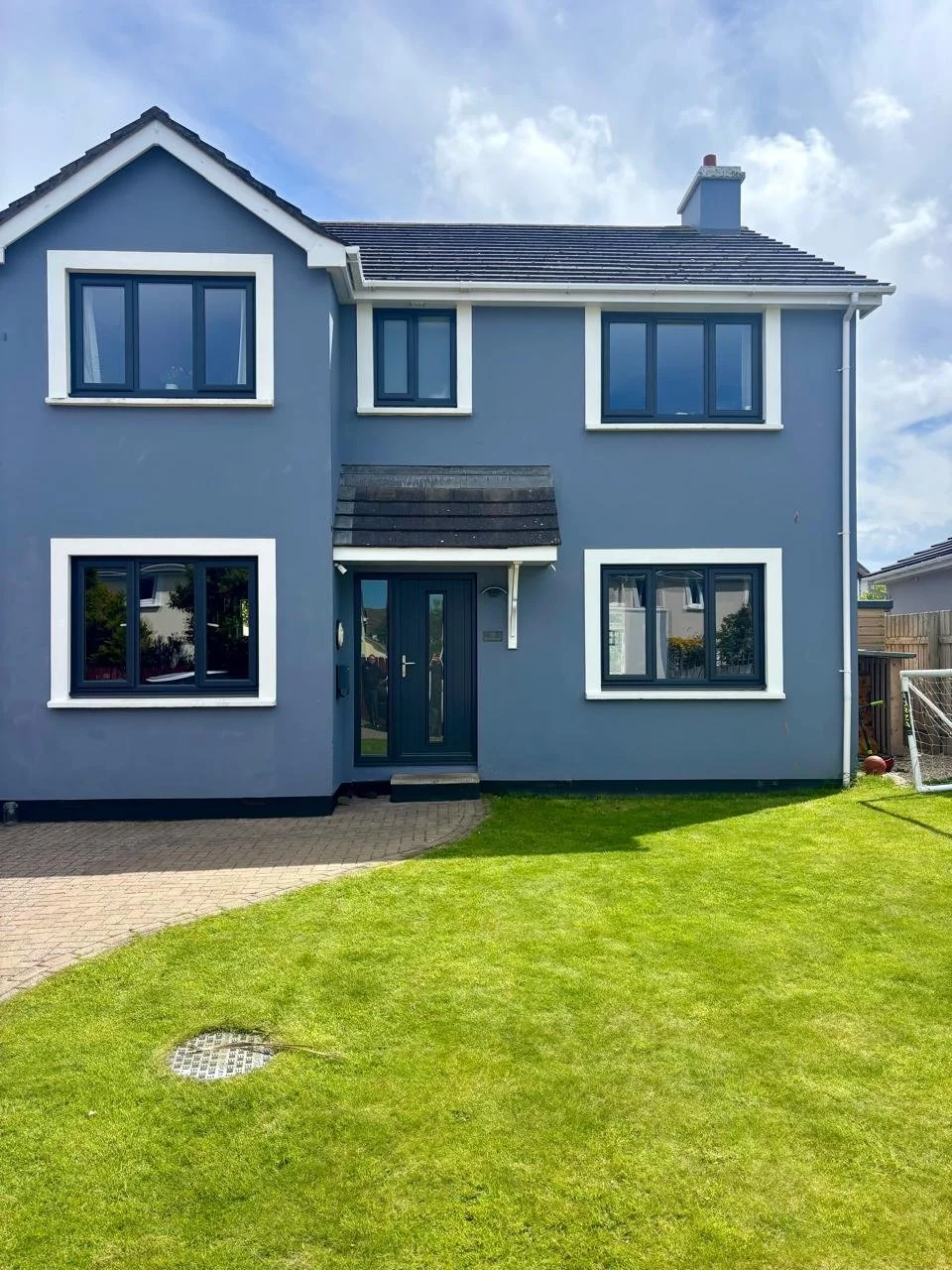 Front view of a gray two-story house with white window frames and a black front door, surrounded by a well-maintained green lawn and a paved pathway.