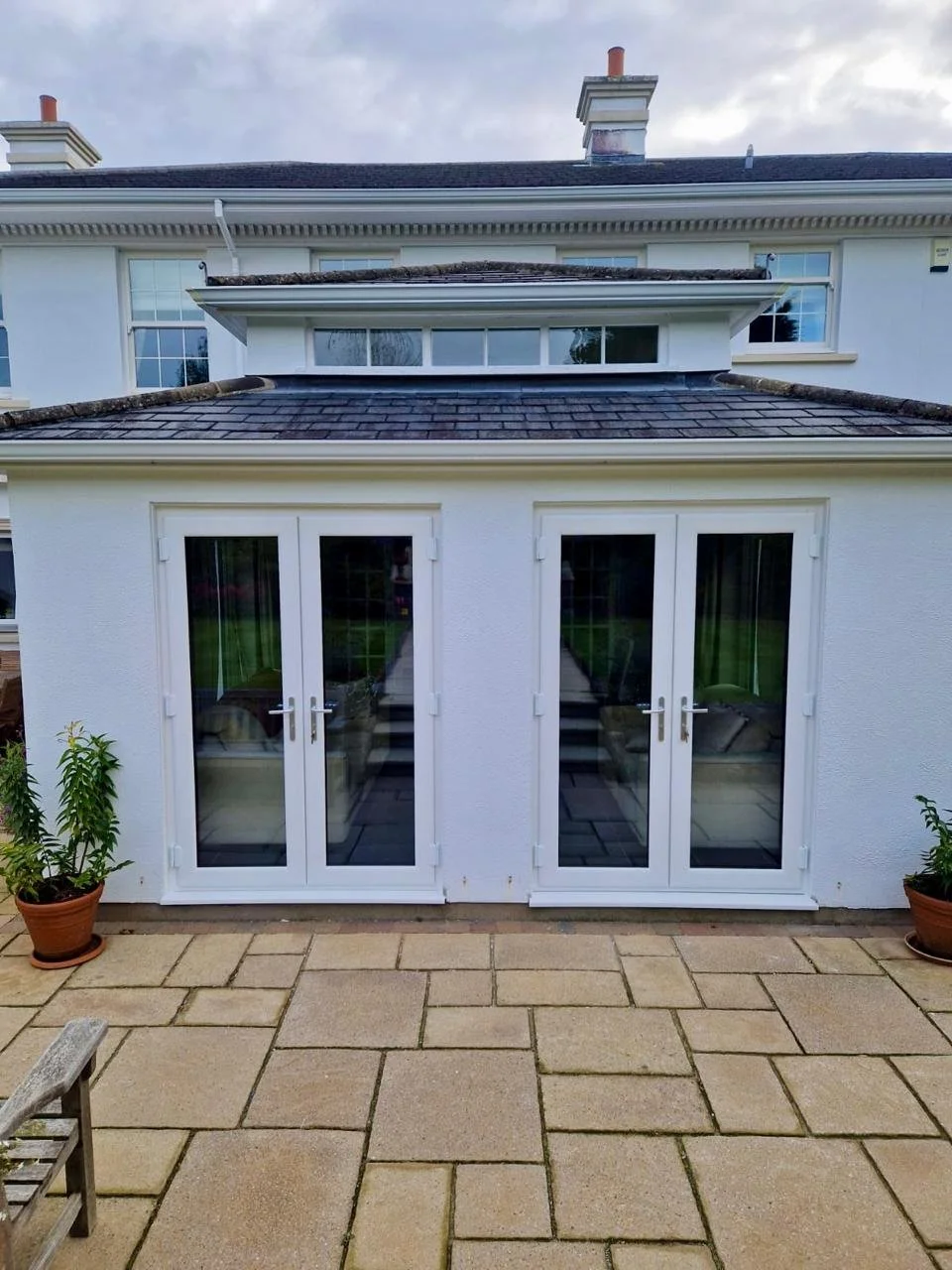 White house with large glass double doors on the patio, potted plants on each side, and a brick-patterned patio in foreground.