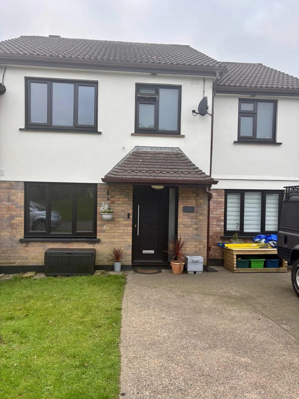 Front of a modern two-story house with a brick and white exterior, black front door, grey window frames, and a small front yard with a driveway and garden plants.