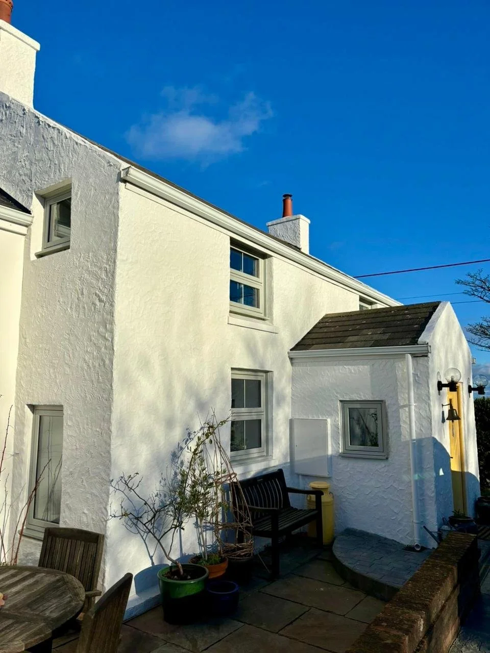 White house exterior with textured walls, several windows, a wooden bench, potted plants, an outdoor light fixture, and a small paved patio area under a clear blue sky.