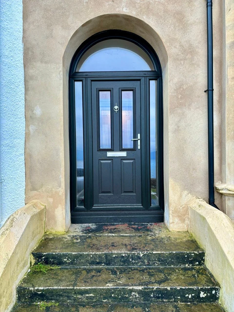 Black front door with glass panels, surrounded by an arched frame, on a weathered stone porch with moss and dirt, in a beige stucco house.