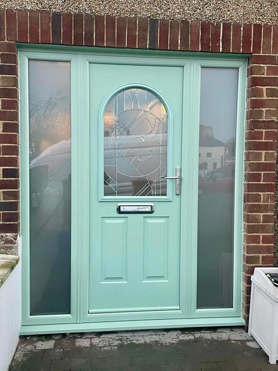 Light green front door with decorative glass panel and sidelights, set in a brick wall, with a silver handle and letterbox.