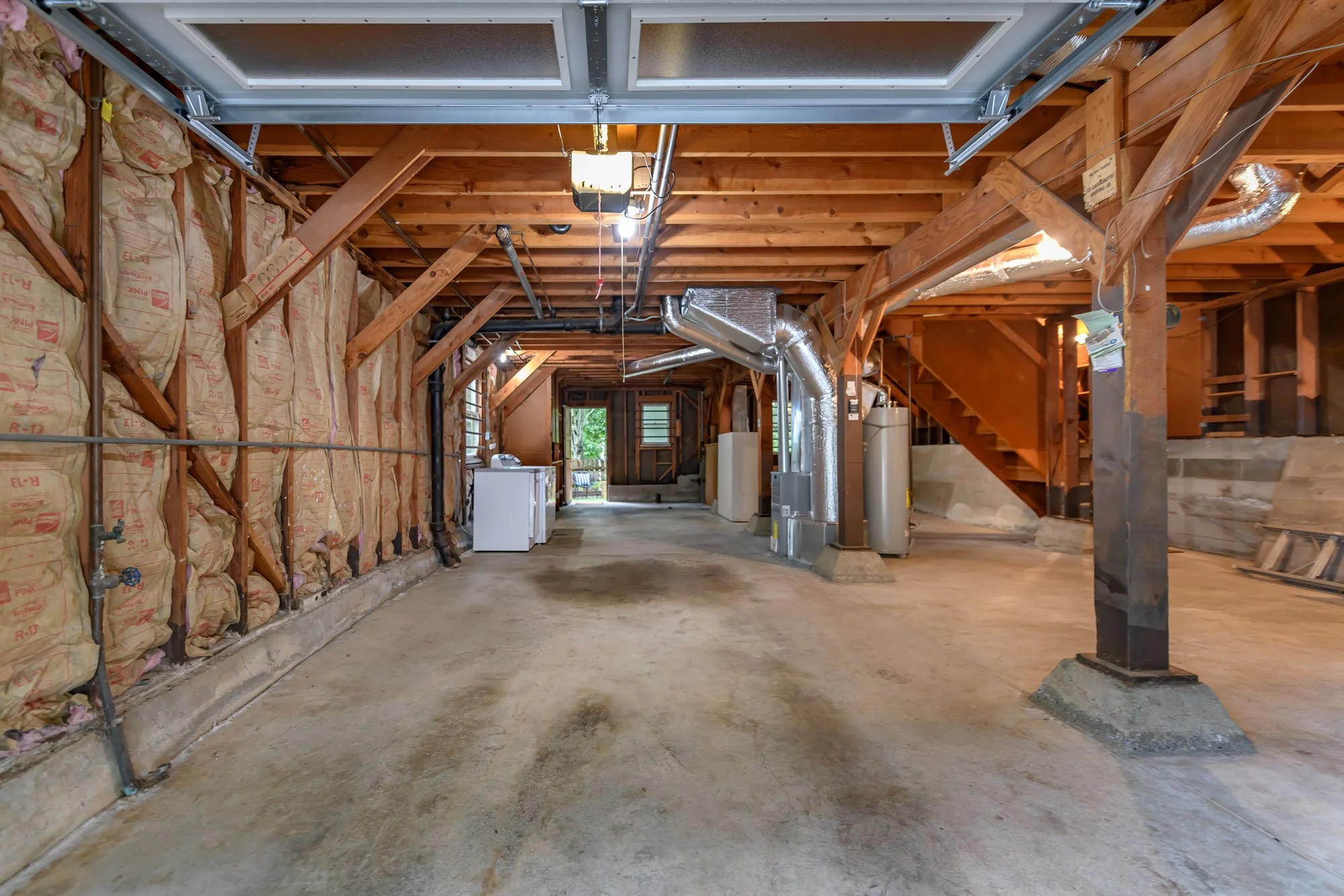 An unfinished basement with visible insulation, plumbing, and ductwork. Laundry appliances and a door leading outside are visible at the far end.