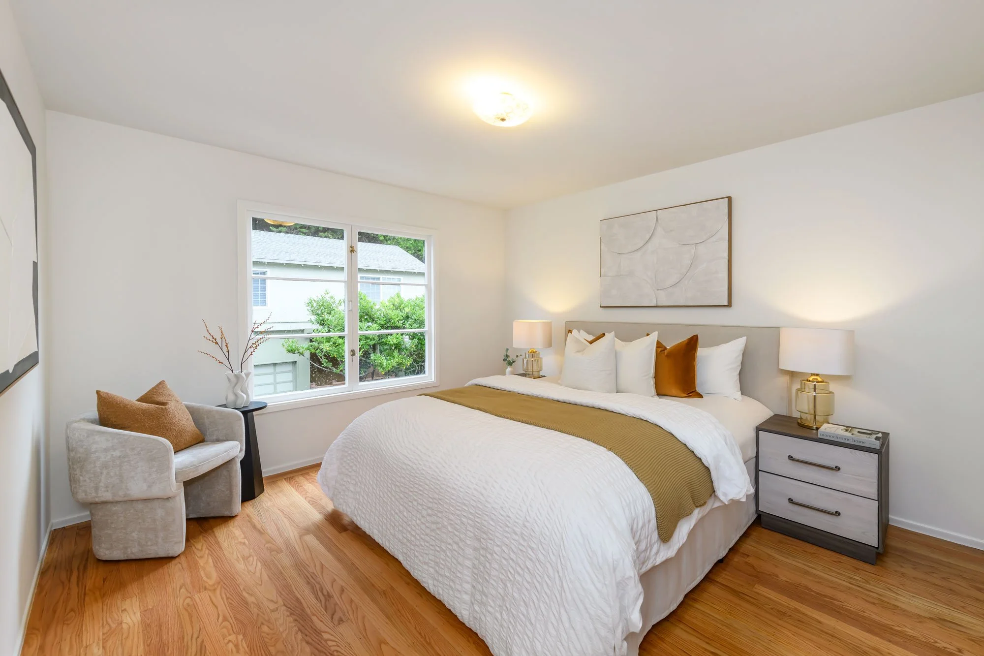Bedroom with white walls, hardwood floor, large bed with white bedding and brown and white pillows, gray nightstand with a lamp, small armchair with a brown pillow, window with a view of greenery and neighboring house, abstract wall art above the bed