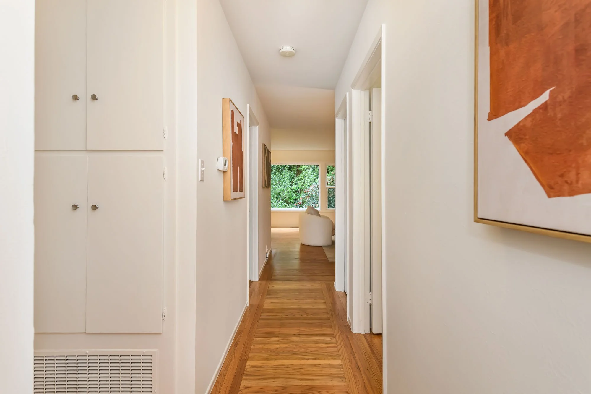 Empty hallway in a house with white walls, wooden flooring, and art on the walls, leading to a brighter room with large windows and greenery outside.