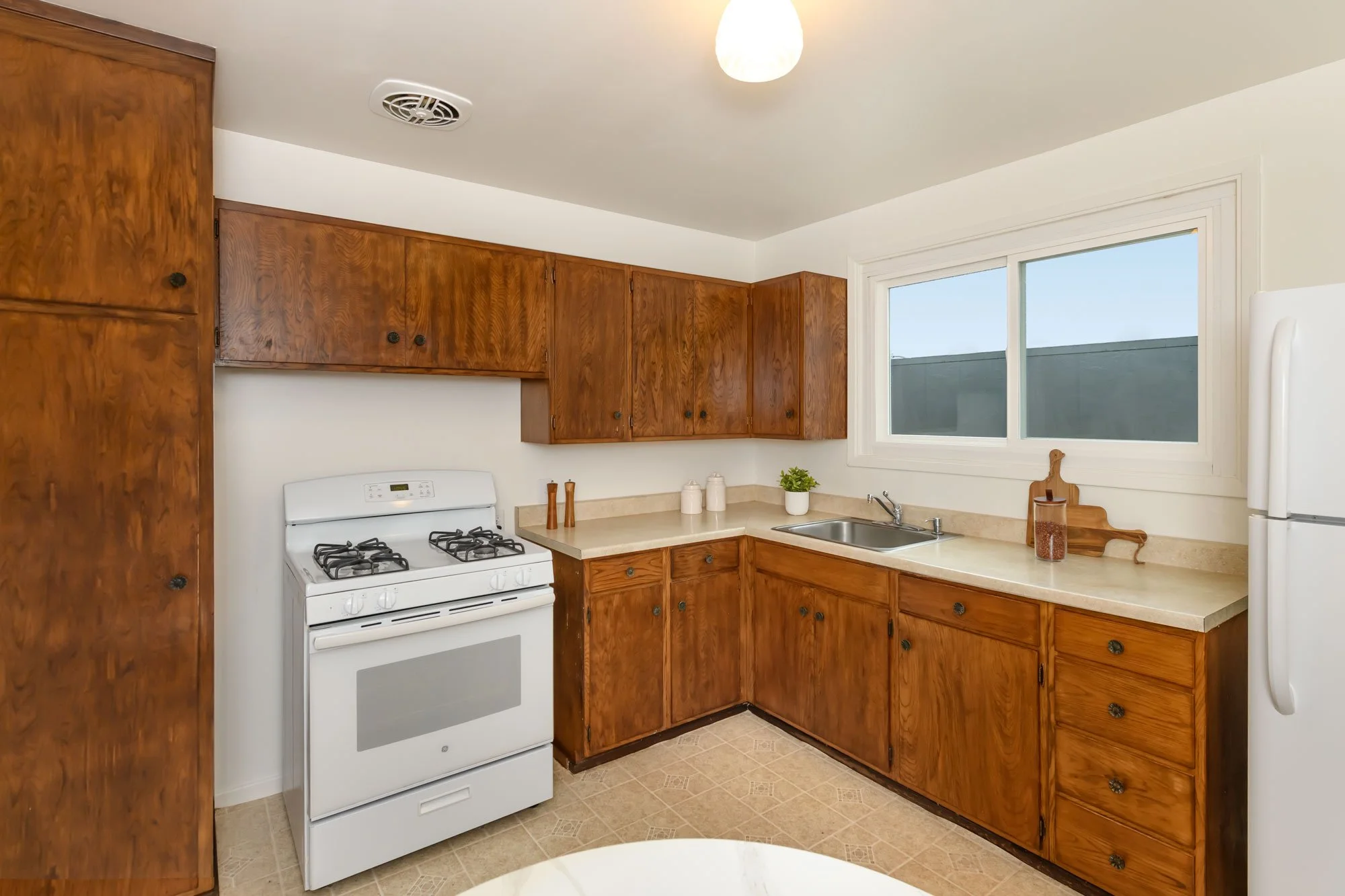 Kitchen with wooden cabinets, white stove, white refrigerator, beige countertops, and a window above the sink.