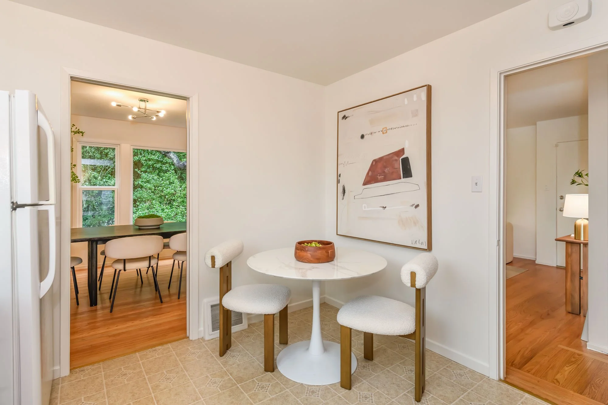 Modern dining area with a white marble round table and two matching chairs, wall art, and a bowl of fruit, adjacent to a kitchen and a hallway.
