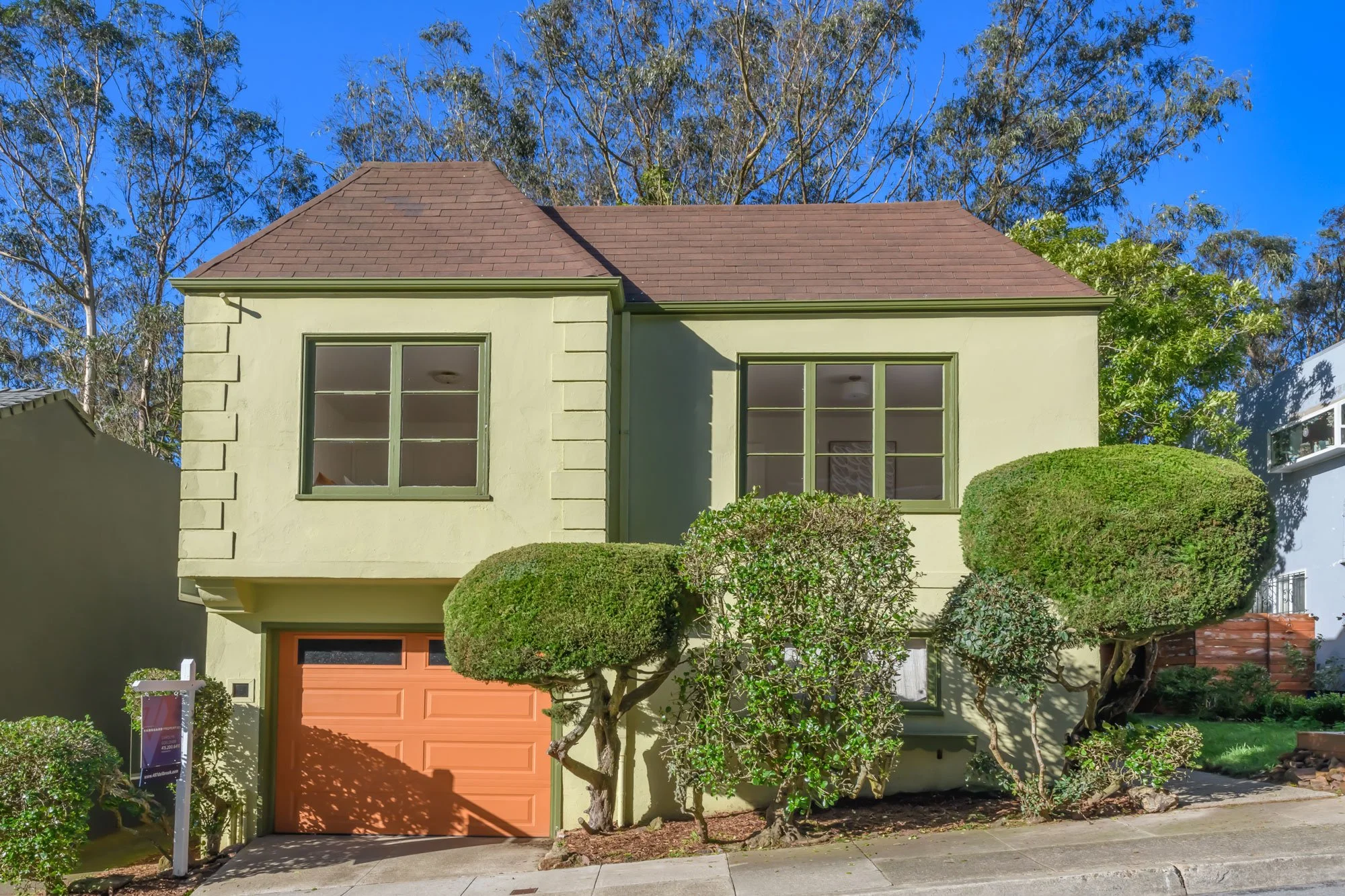 A two-story house with a light green exterior, brown roof, and green window frames. The house has a garage with a peach-colored door and well-maintained bushes in the front yard.