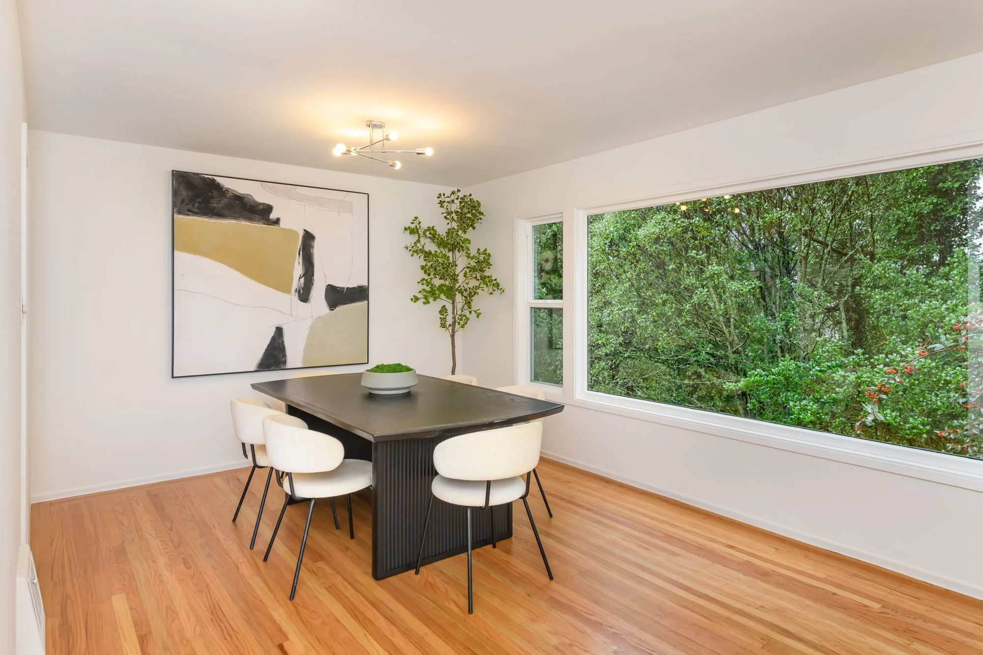 Modern dining room with a black table, six white chairs, a large abstract painting on the wall, a potted plant, and large windows showing greenery outside.