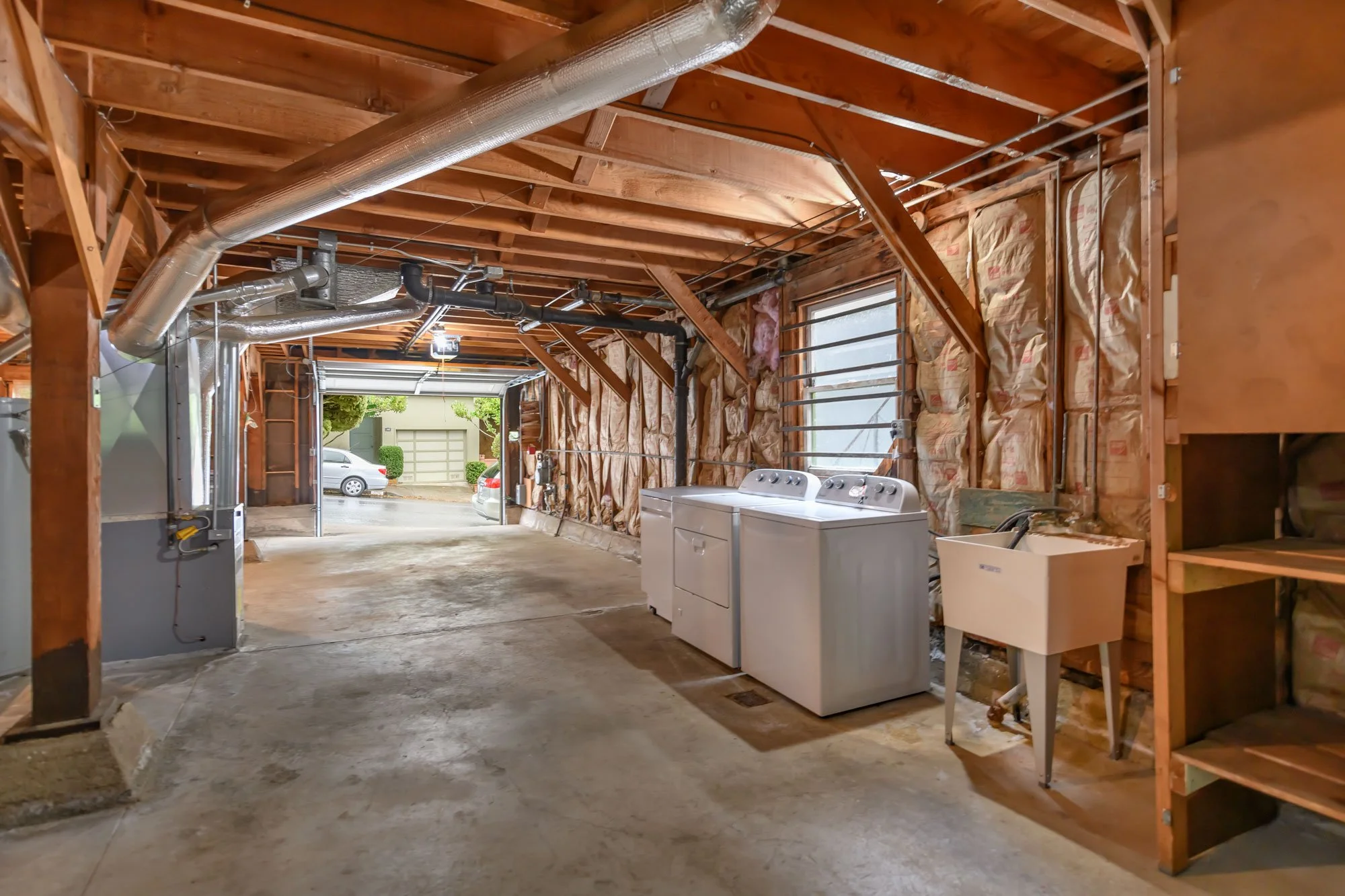An empty garage with exposed wooden beams, insulation on the walls, and a concrete floor. There are laundry appliances, a utility sink, and some storage shelves. The garage door is open, showing a driveway and parked cars outside.