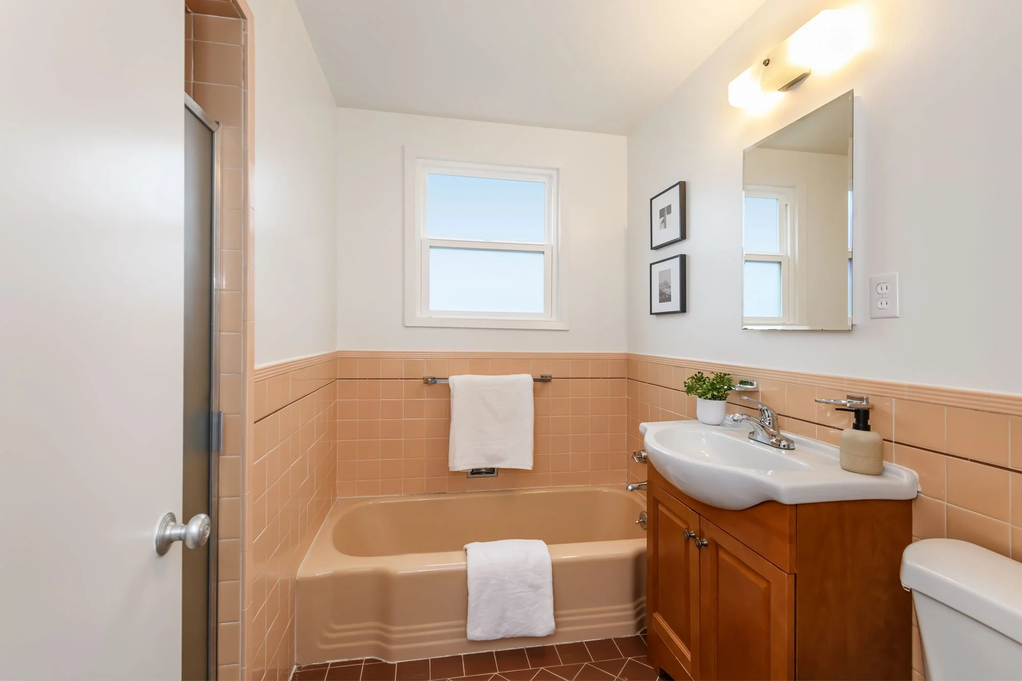 A small bathroom with a peach-colored bathtub, peach tile wainscoting, and white upper walls. There are two small black-and-white framed pictures on the wall, a window above the tub, a wooden vanity with a white sink, a mirror, a potted plant, soap d