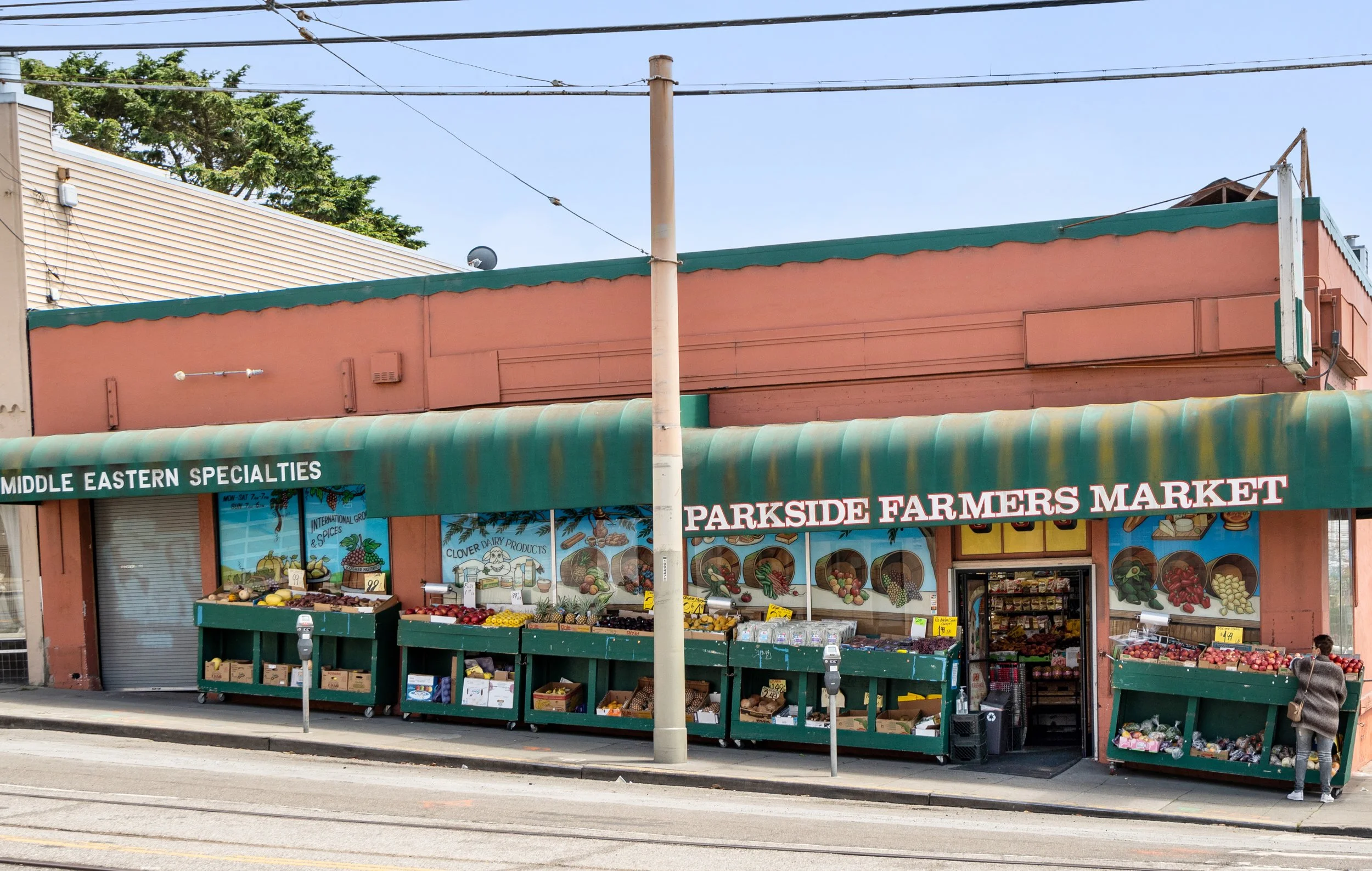 A storefront of Parkside Farmers Market with outdoor produce displays and a green awning, pink building, and a street with tram tracks in front.