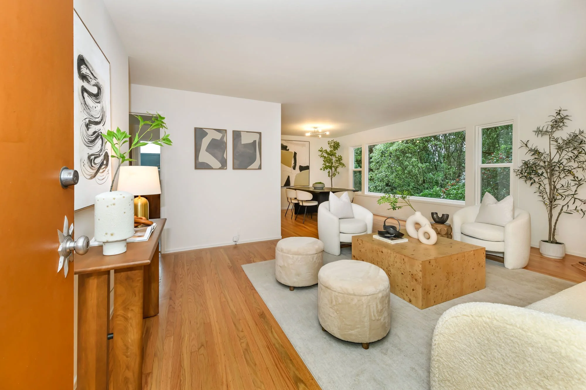 Living room with white armchairs, a wooden coffee table, beige ottomans, and large windows overlooking trees.