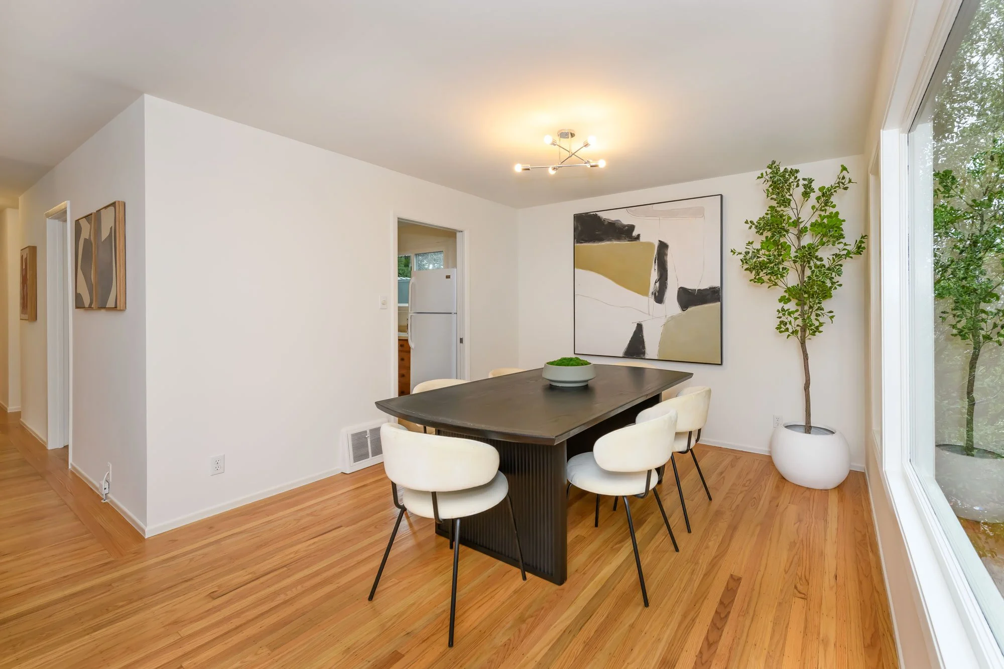 Dining room with a black table, six white chairs, abstract wall art, a large window, and a potted tree.