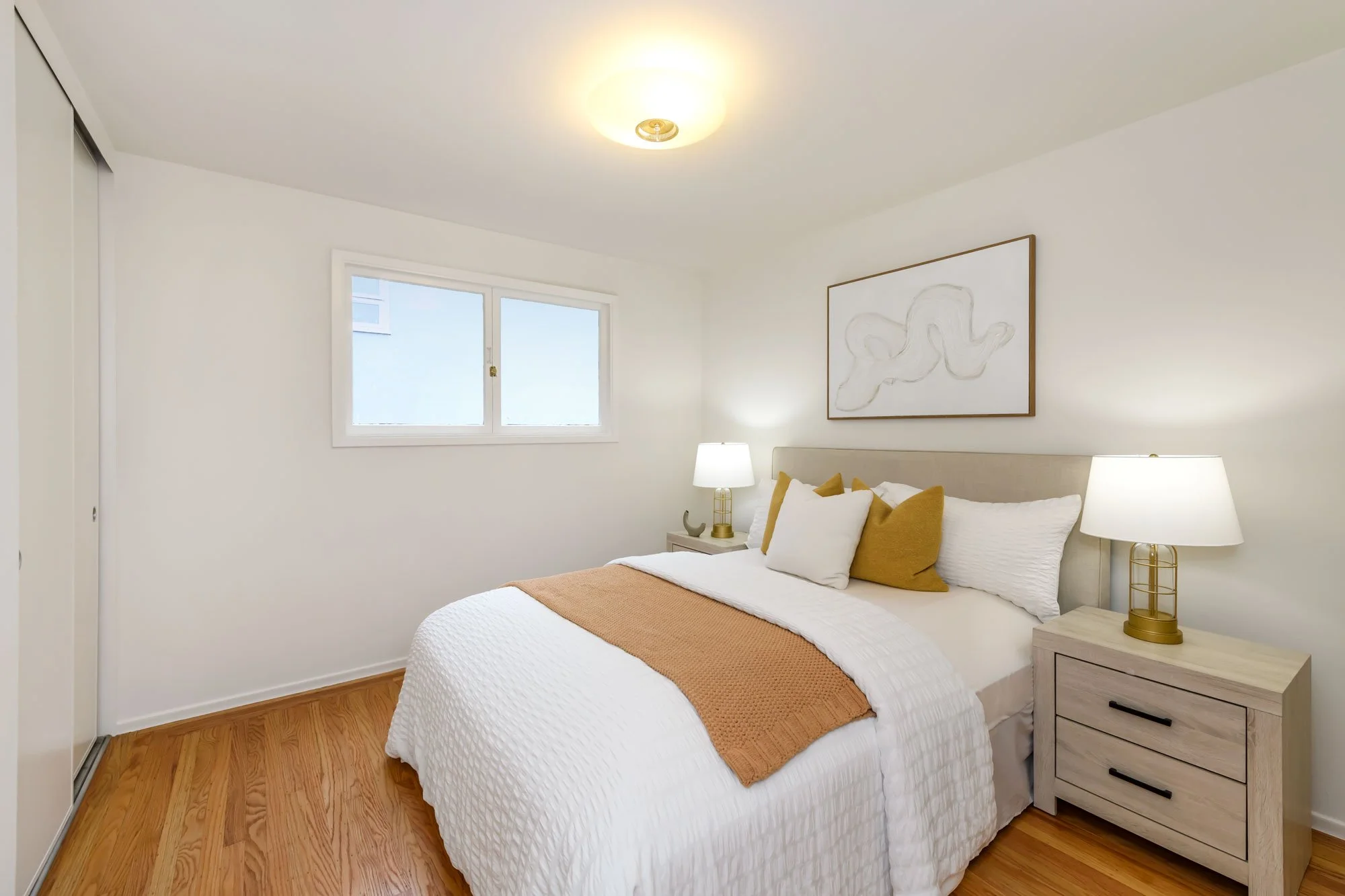 Cozy bedroom with white walls, a bed with white bedding, and mustard yellow and white pillows. It has wooden nightstands with matching lamps, a window, and a framed abstract art piece above the bed. Hardwood floors and simple decor.
