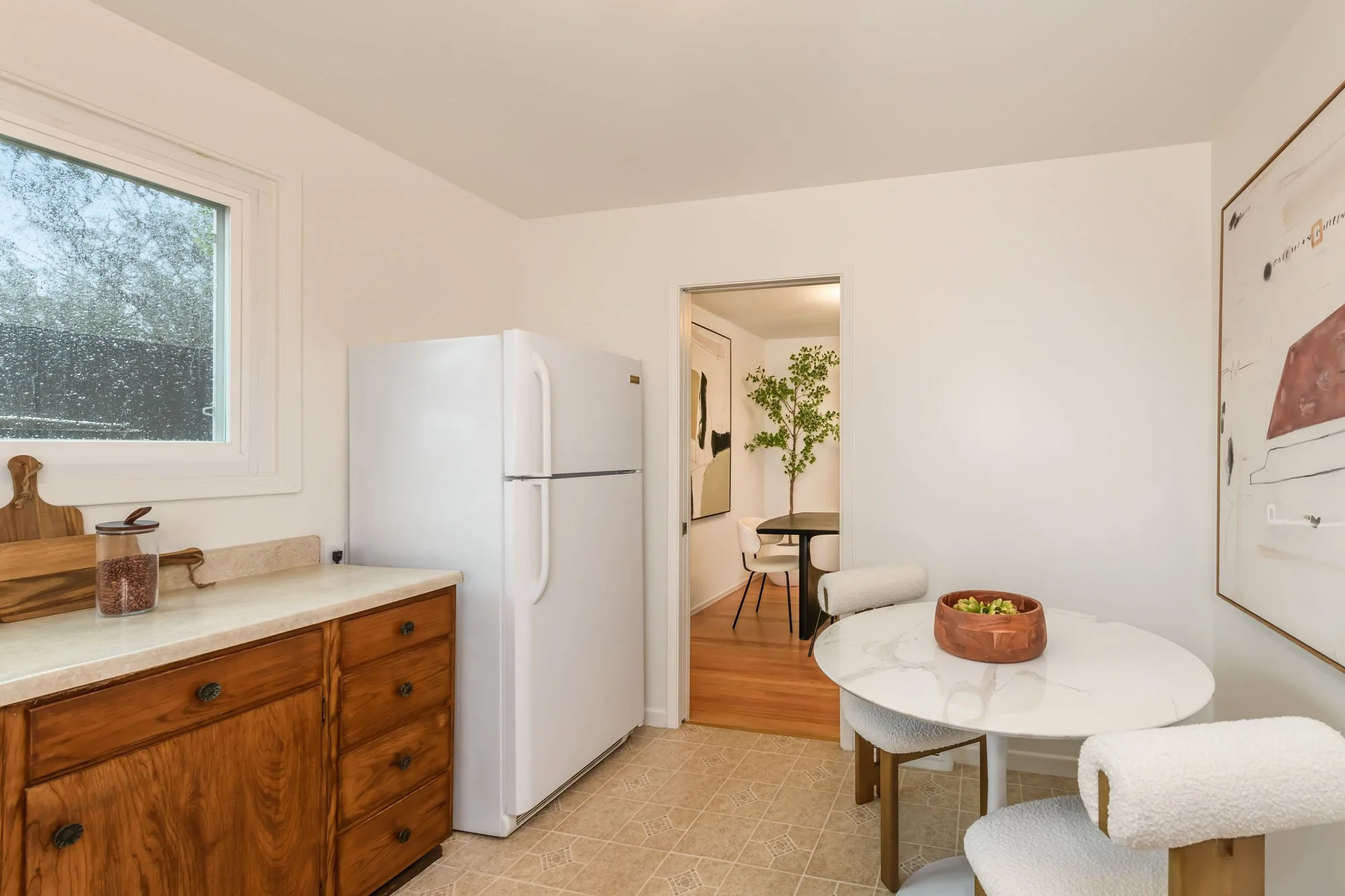 A kitchen with a window, wooden cabinets, a white refrigerator, a round marble table with white chairs, and a bowl of succulents. There is a doorway leading to a dining area with a black table, white chairs, and a large potted plant.