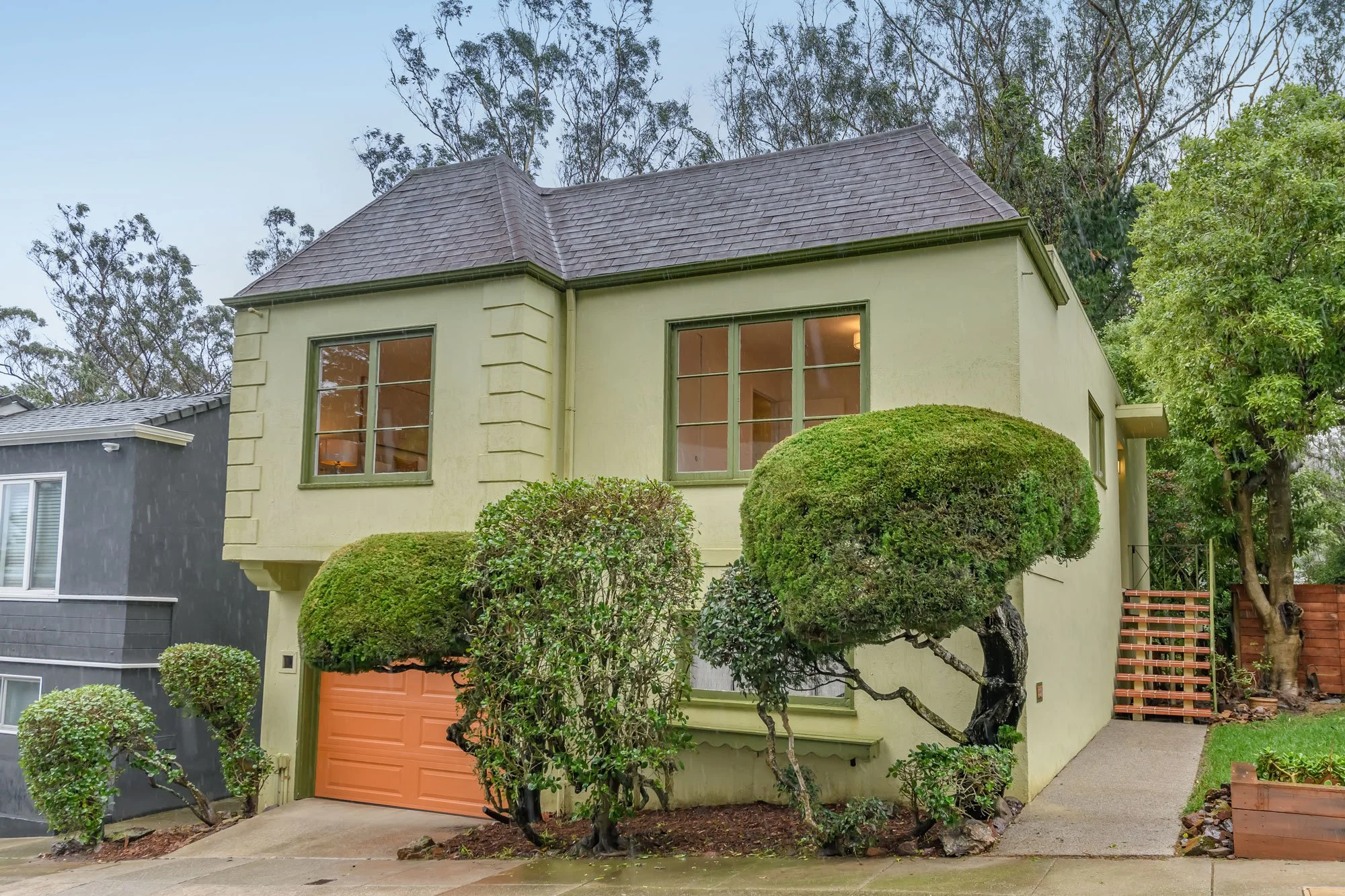 A two-story house with pale yellow exterior walls, green window frames, and a light orange garage door. Well-maintained shrubs and trees are in the front yard, with a sidewalk leading to the side of the house. The sky is overcast, and neighboring hou
