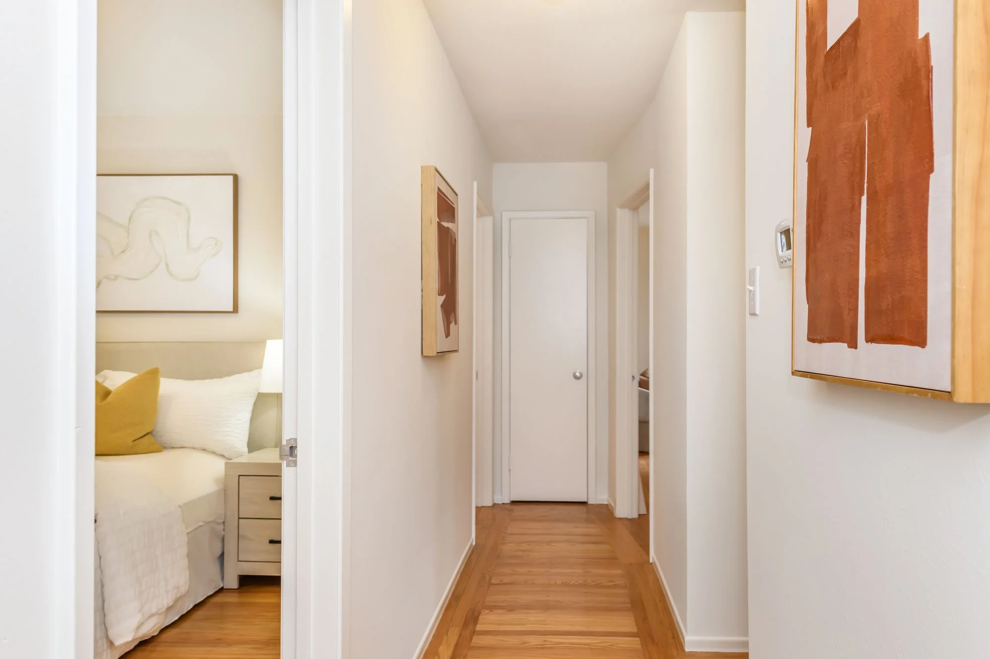 Hallway with hardwood floor, leading to closed door, with artwork on walls, and an open doorway to a bedroom visible on the left.