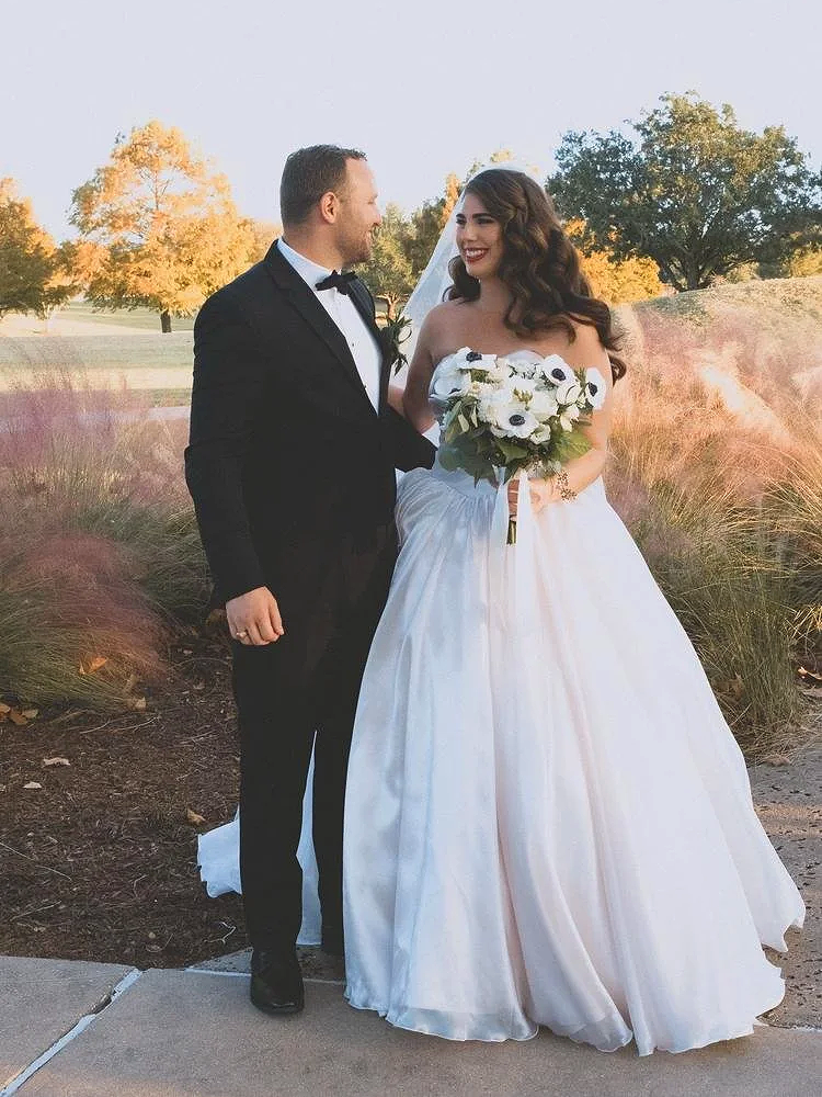 A bride and groom standing outdoors during sunset, dressed in wedding attire, smiling at each other. The bride wears a strapless white gown and holds a bouquet of white flowers. The groom is in a black tuxedo with a bow tie and boutonniere. The background features trees with autumn foliage and pink ornamental grass.